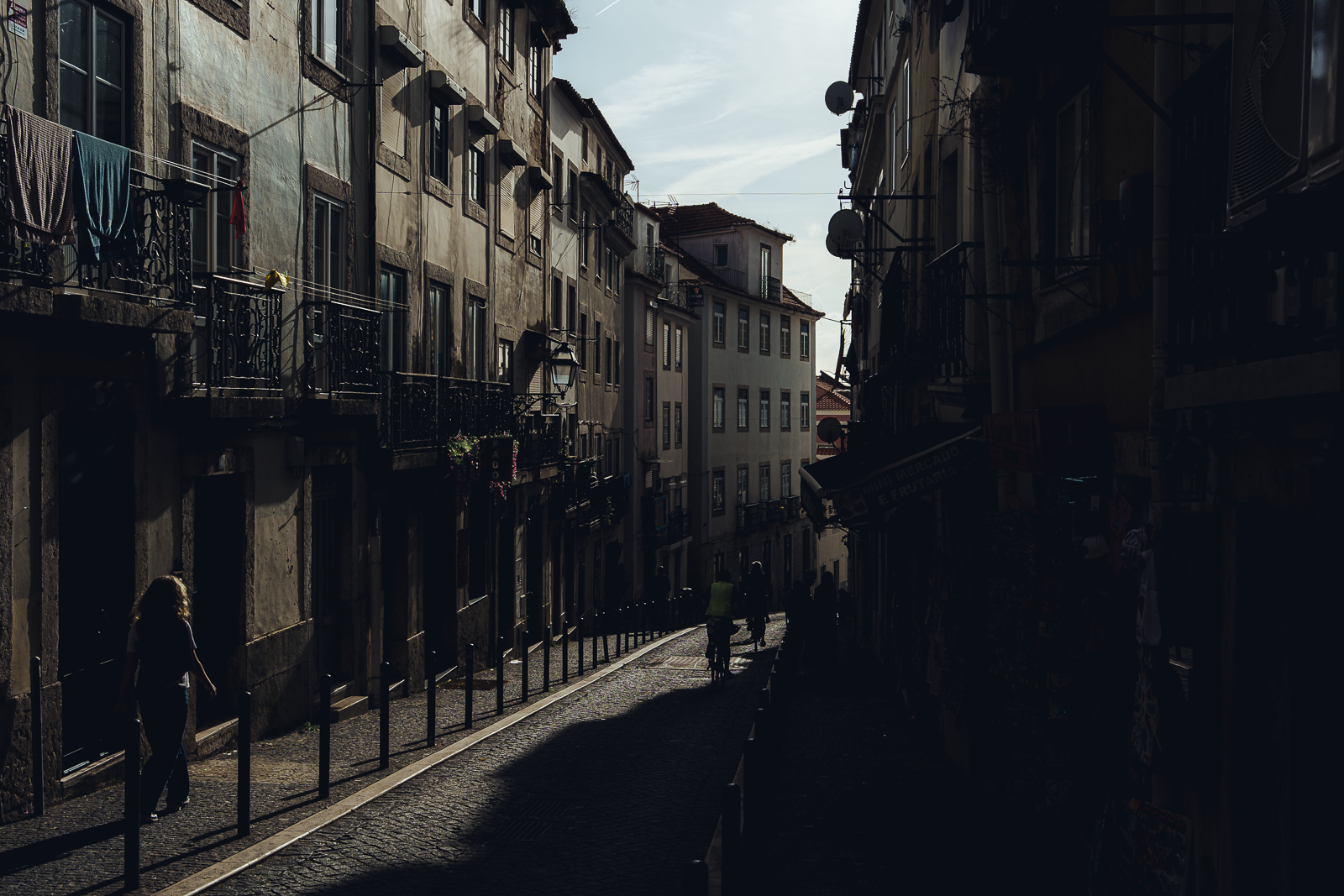 A narrow urban street with old apartment buildings on either side, some with laundry hanging on balconies, and a few pedestrians walking in the shadows with sunlight illuminating the background sky.