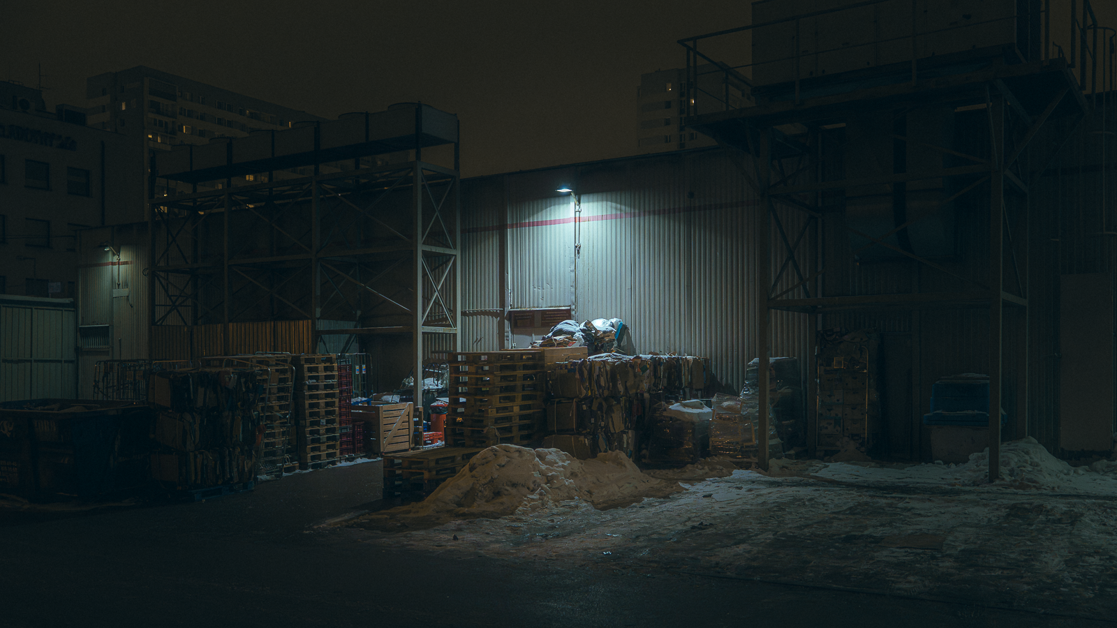 An industrial outdoor storage area at night with pallets, stacked items, and snow with a metal building and high-rise buildings in the background, illuminated by a single overhead light.