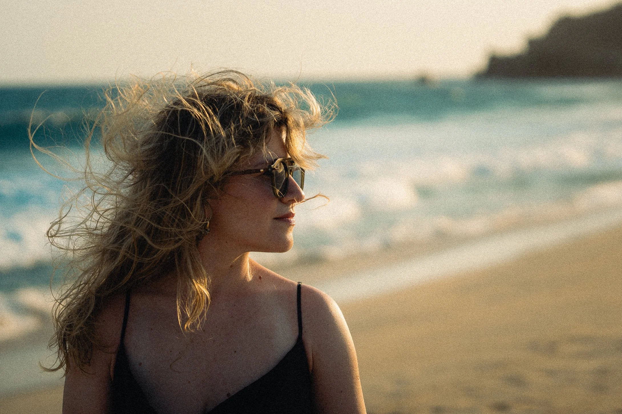 A woman with curly blonde hair wearing sunglasses and a black spaghetti strap top standing on a sandy beach with ocean waves and a distant landmass in the background during sunset.