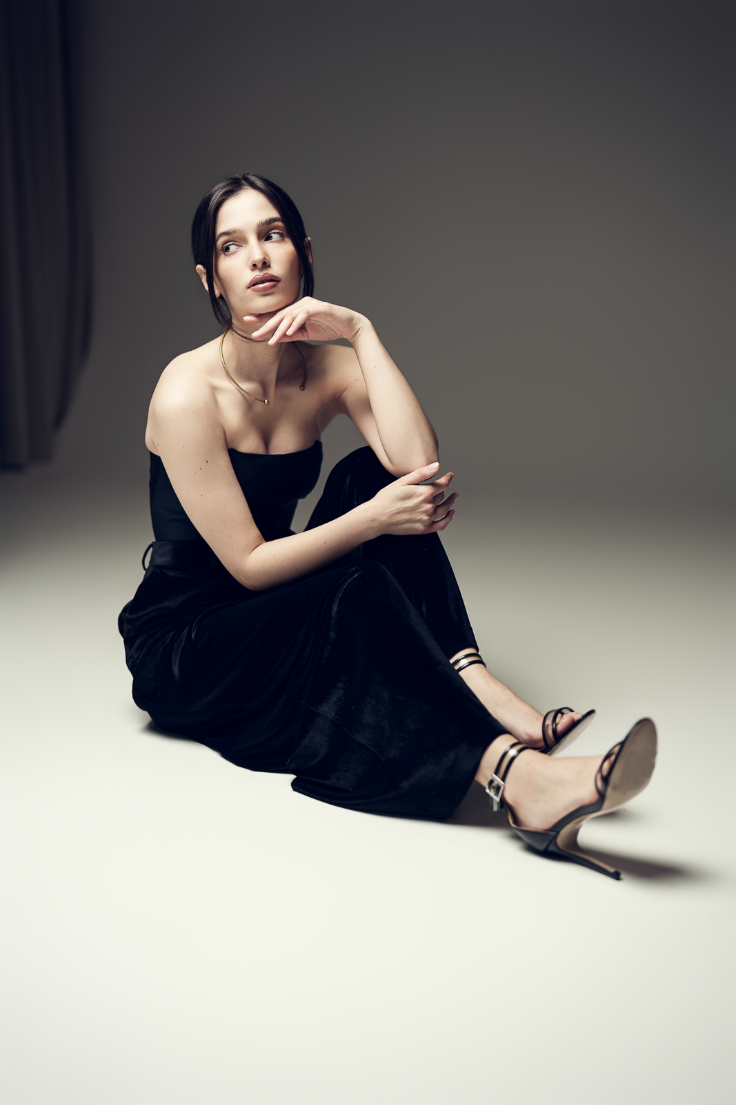 A woman sitting on the floor in a black strapless dress and high heels, looking to the side with her hand near her chin, in a minimalistic studio setting.