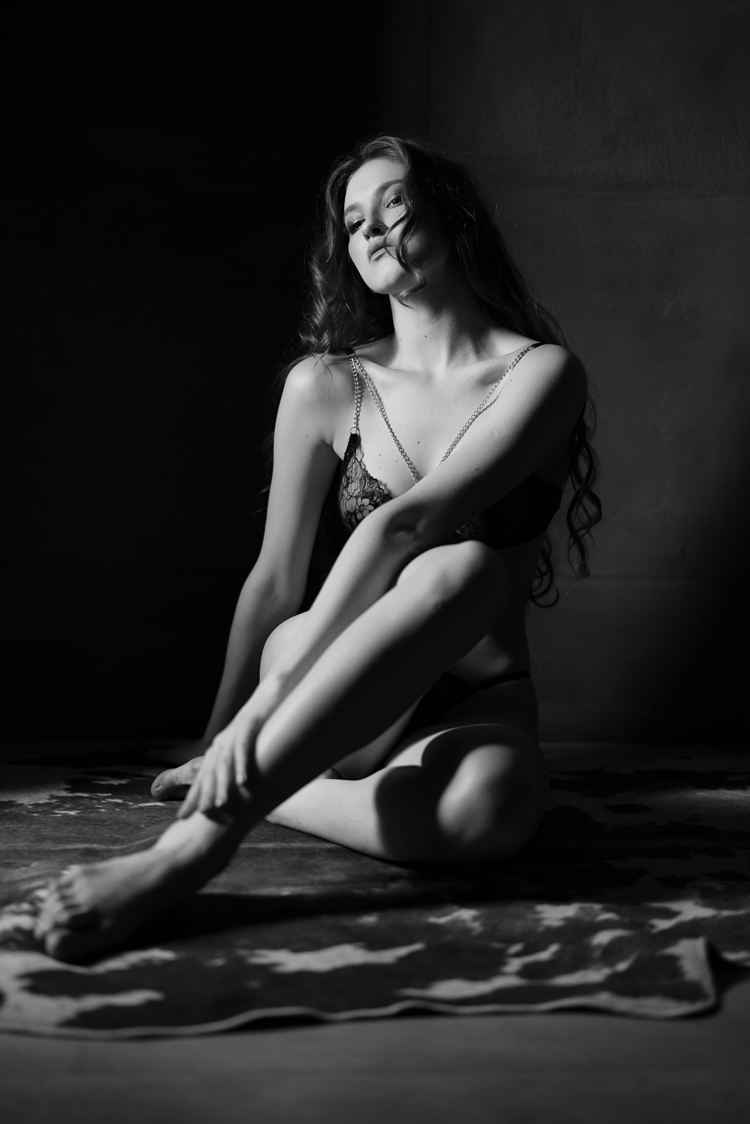 A woman with long curly hair sitting on the floor with her knees bent, wearing lingerie and chains, in black and white.