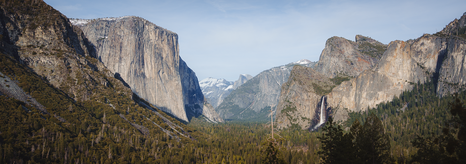 A panoramic view of Yosemite Valley with granite cliffs, lush green forests, and a waterfall cascading down a mountain.