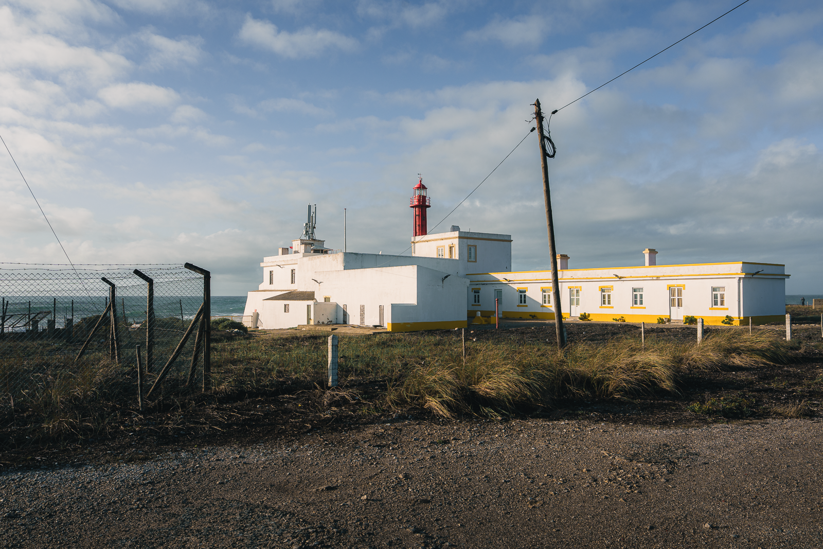 A lighthouse with a white building and red tower against a partly cloudy sky, fenced in with a dirt road in the foreground.