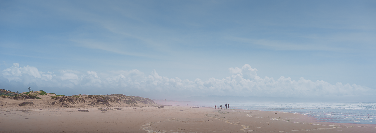 A wide view of a sandy beach with dunes on the left and the ocean on the right. A few people are walking along the shoreline under a partly cloudy sky.