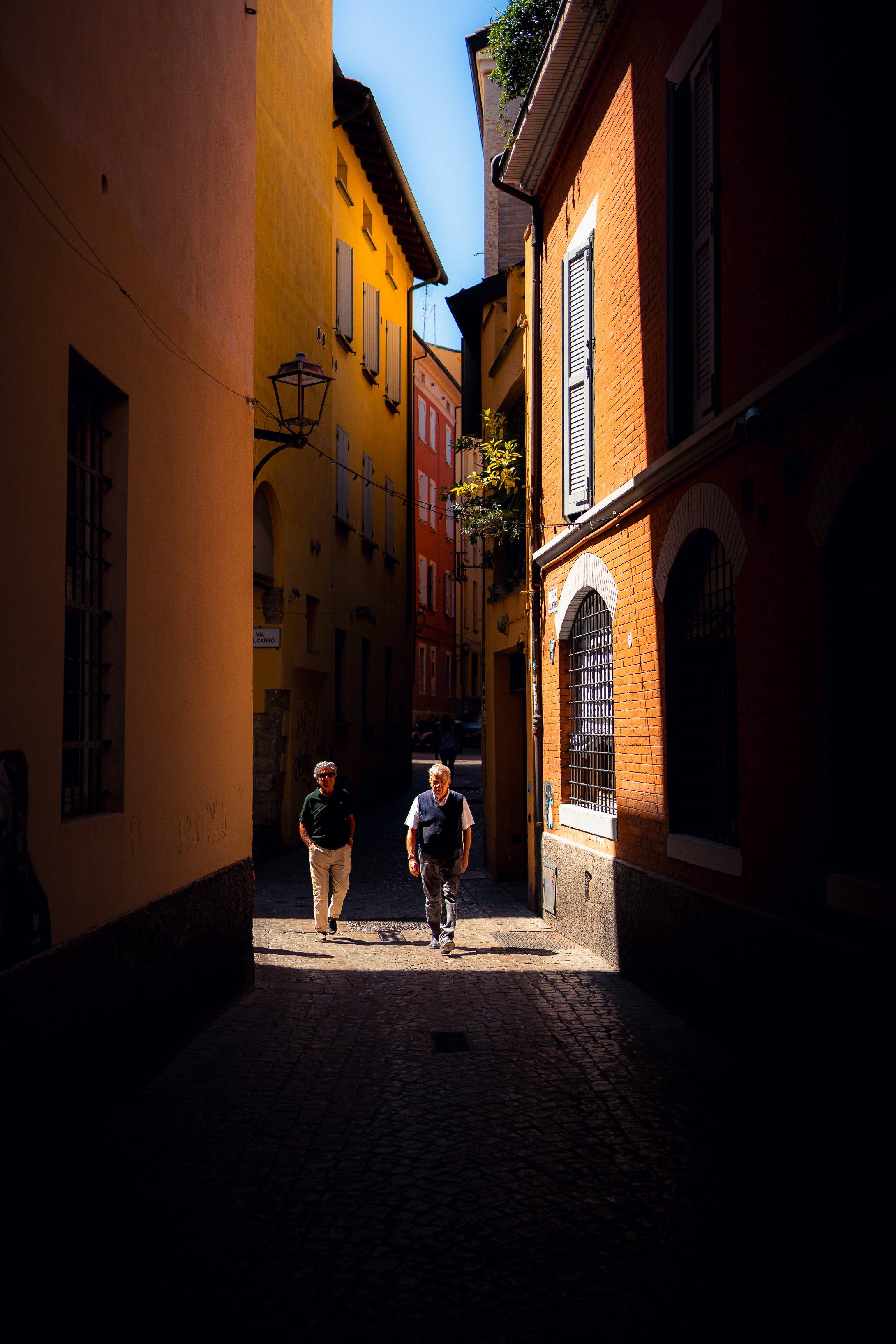Two men walking through a narrow, sunlit alleyway lined with colorful buildings, some with shutters and iron window bars.