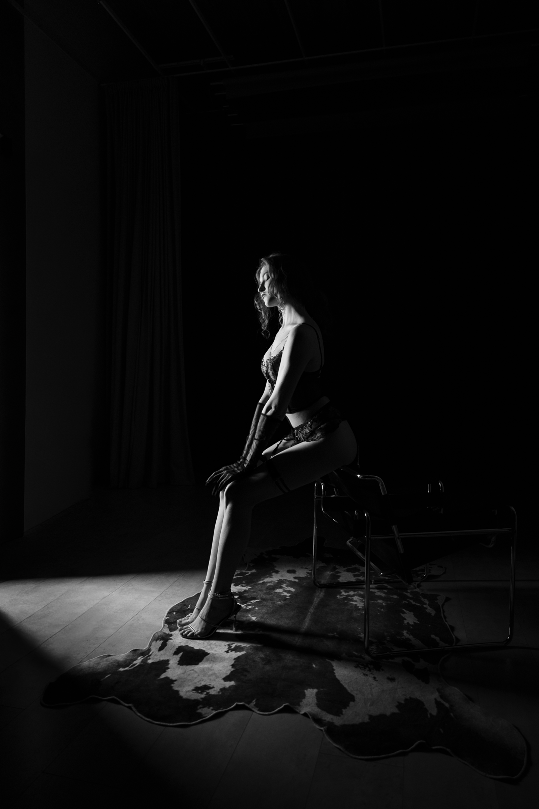 A black and white photo of a woman sitting on a chair in lingerie, with a patterned rug beneath her and a dark background.