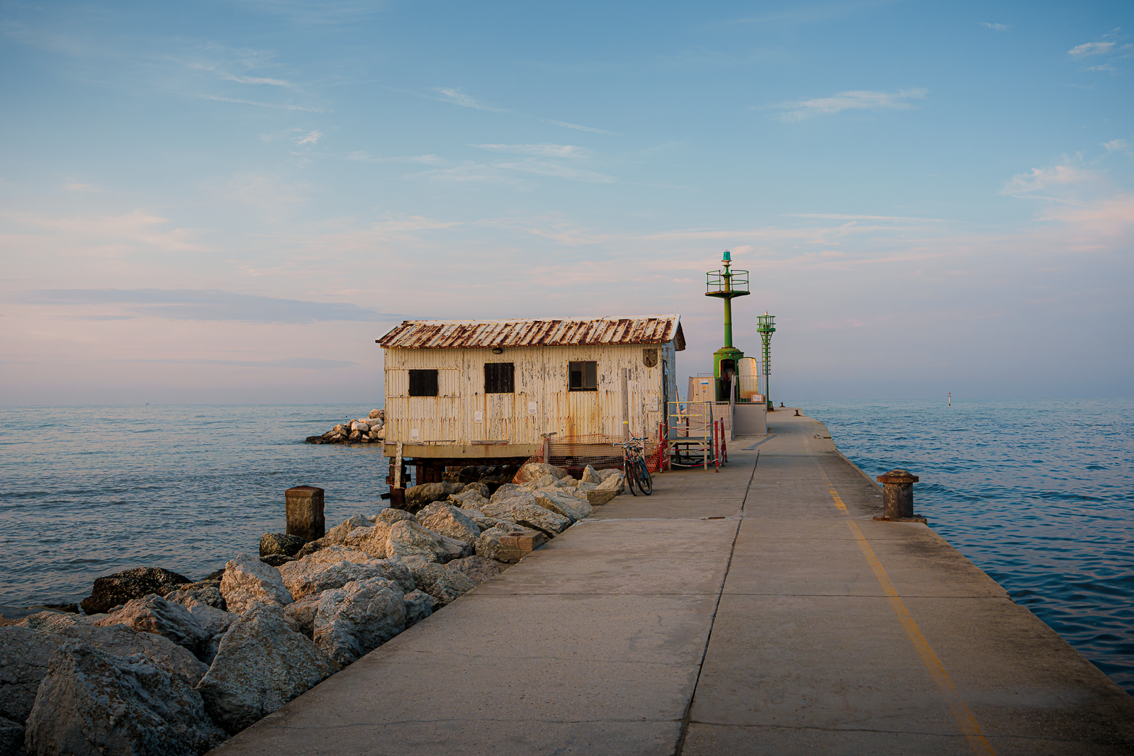 A weathered wooden hut on a pier over the water with a bicycle parked nearby and a lighthouse in the background, under a partly cloudy sky.