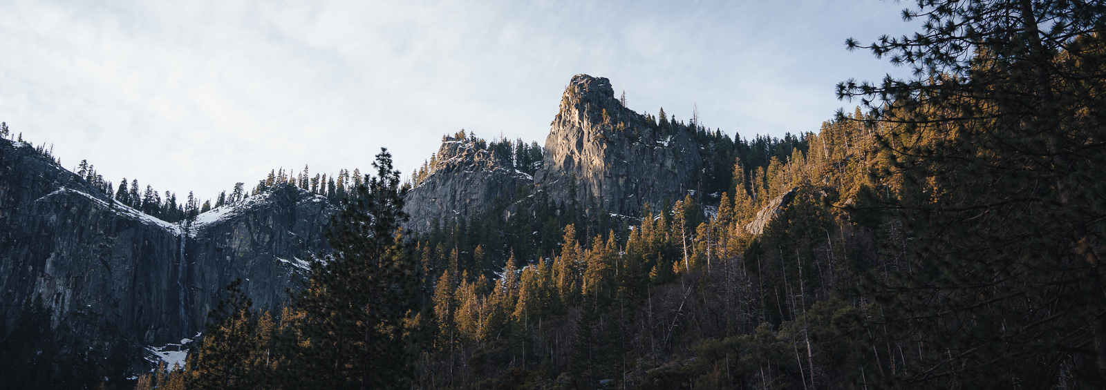 Mountains with rocky cliffs, evergreen trees, and snow patches, under a partly cloudy sky.