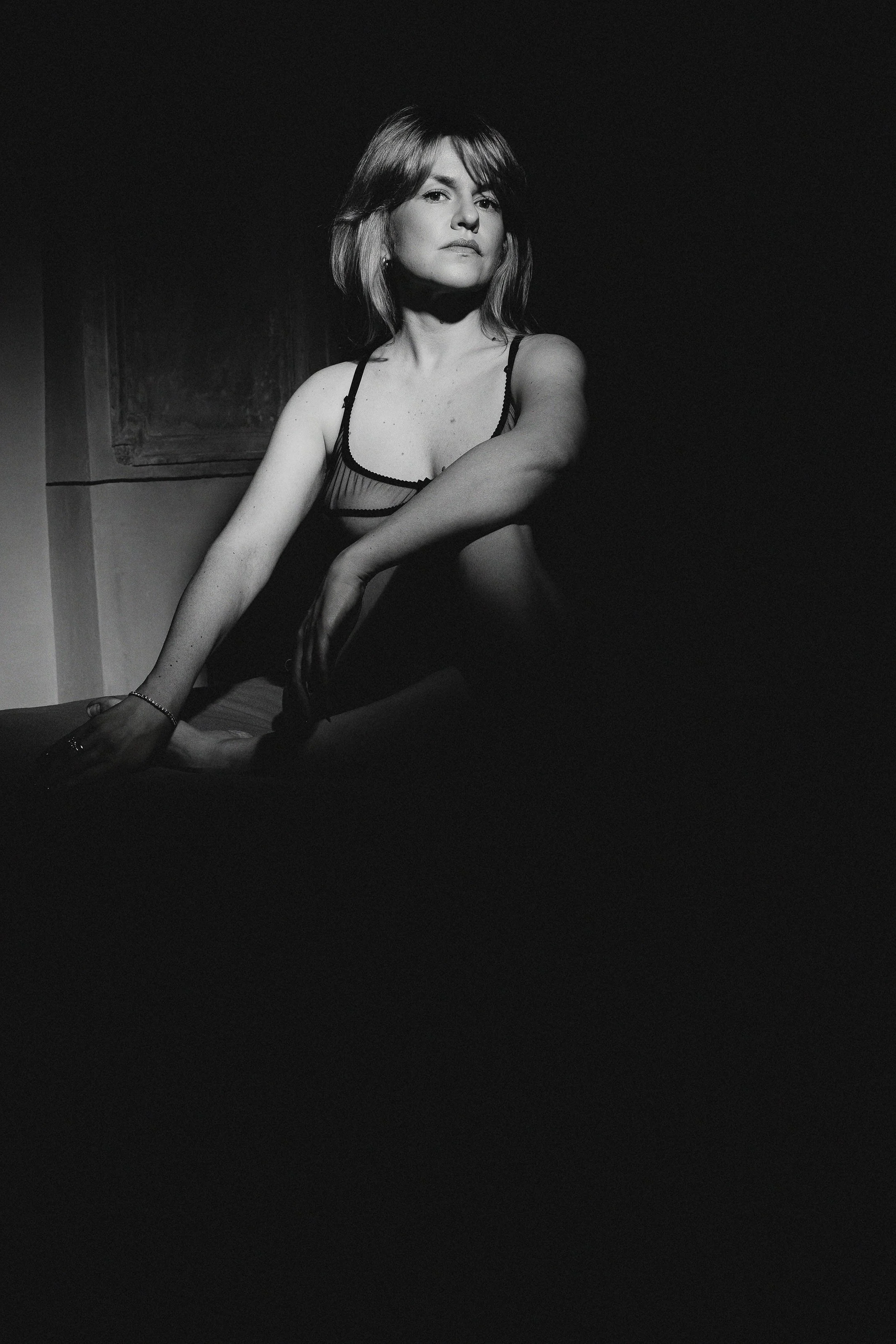 A woman in lingerie sitting on a bed, looking to the side with a serious expression, captured in black and white.