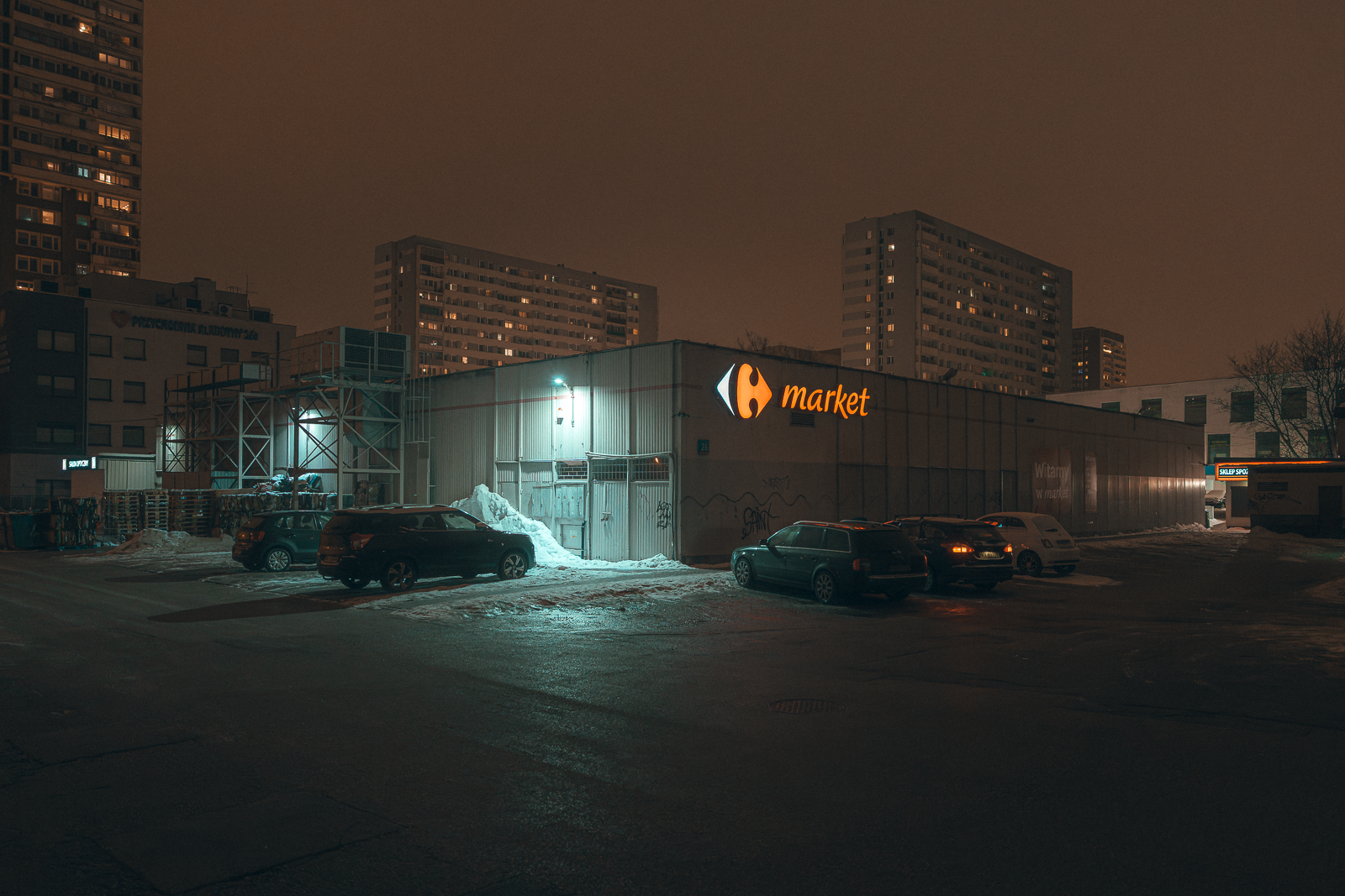 A parking lot outside a grocery store at night, with some cars parked and snow on the ground. The store has a sign that reads 'market' with a logo of an eye, and apartment buildings are visible in the background under a cloudy sky.