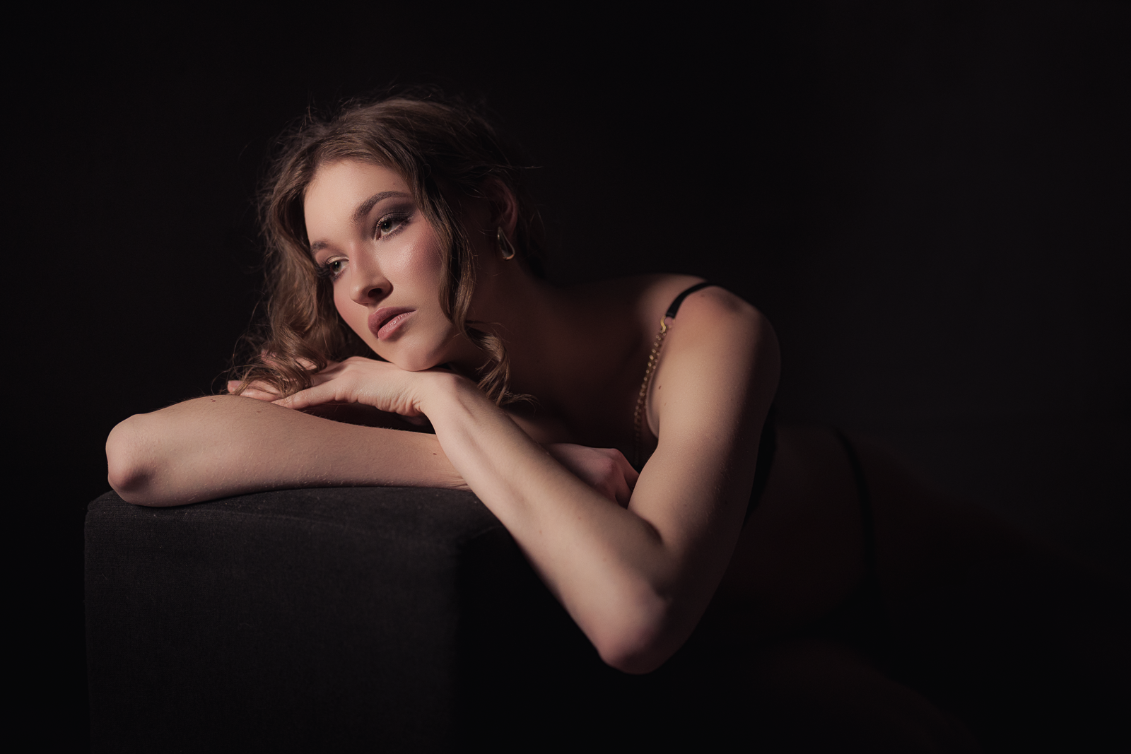 A woman with wavy hair lying on her side, resting her chin on her arm, in a dark setting with soft lighting highlighting her face and shoulder.