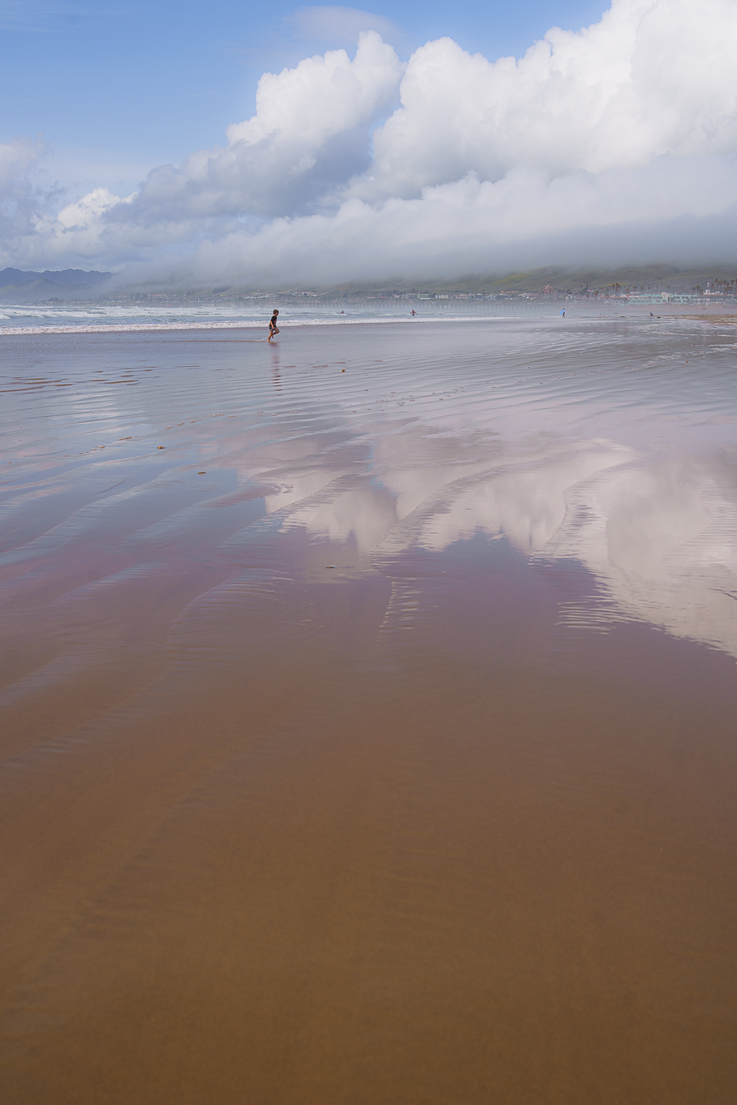 Person walking on a wide, sandy beach with cloudy skies and mountains in the background, with reflections of clouds on the wet sand.