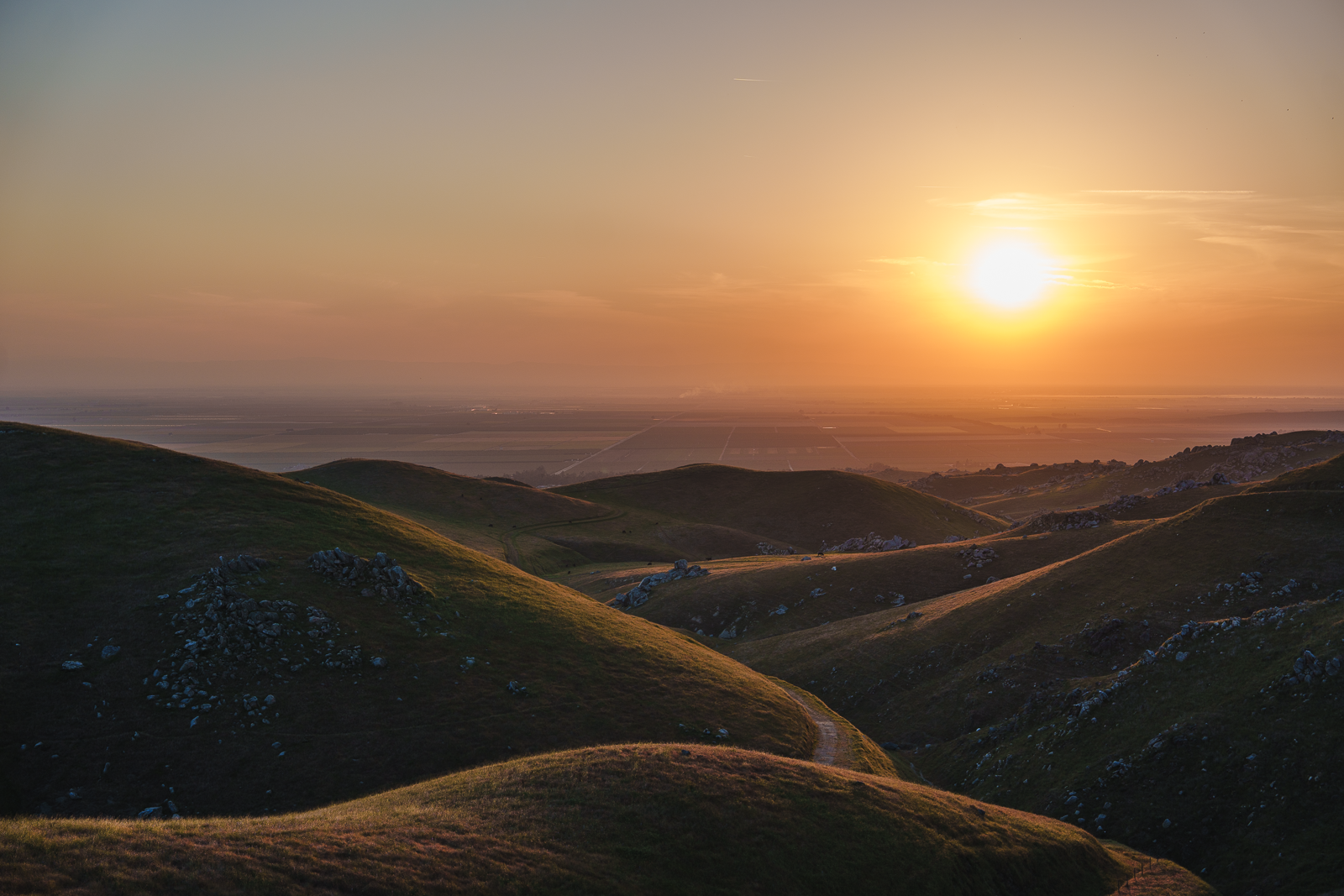 Sunset over rolling green hills with rocky outcroppings, with the sun near the horizon casting a warm glow.