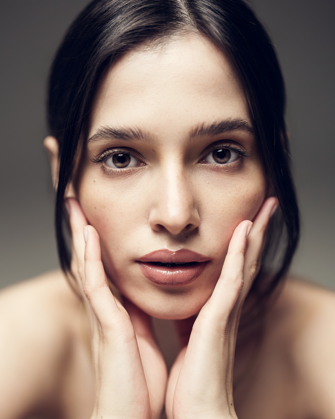 Close-up of a young woman with dark hair, touching her face gently, looking directly at the camera.