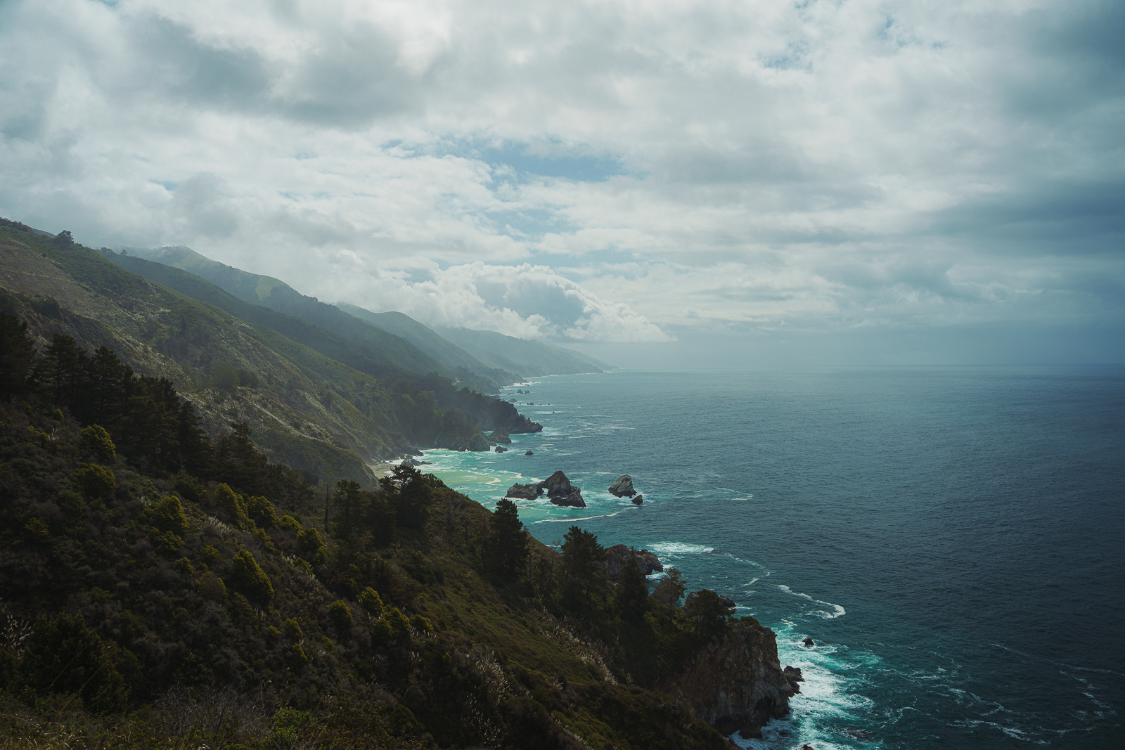 A scenic view of a rugged coastline with green hills and trees descending into the ocean, under a cloudy sky.