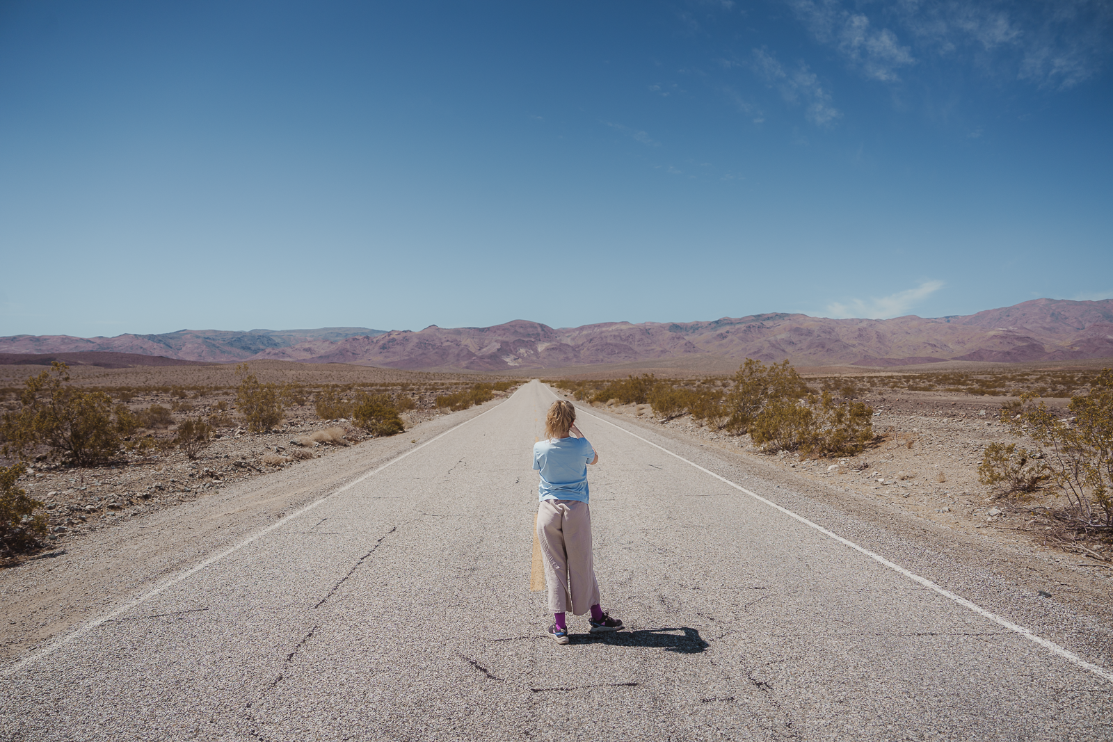 A person standing in the middle of an empty desert road with mountains in the background under a clear blue sky.