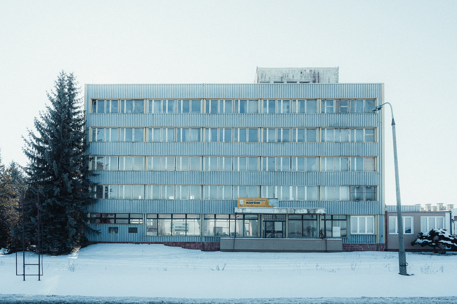 A large multi-story building with numerous windows, a sign at the entrance, and some trees and snow in front of it.