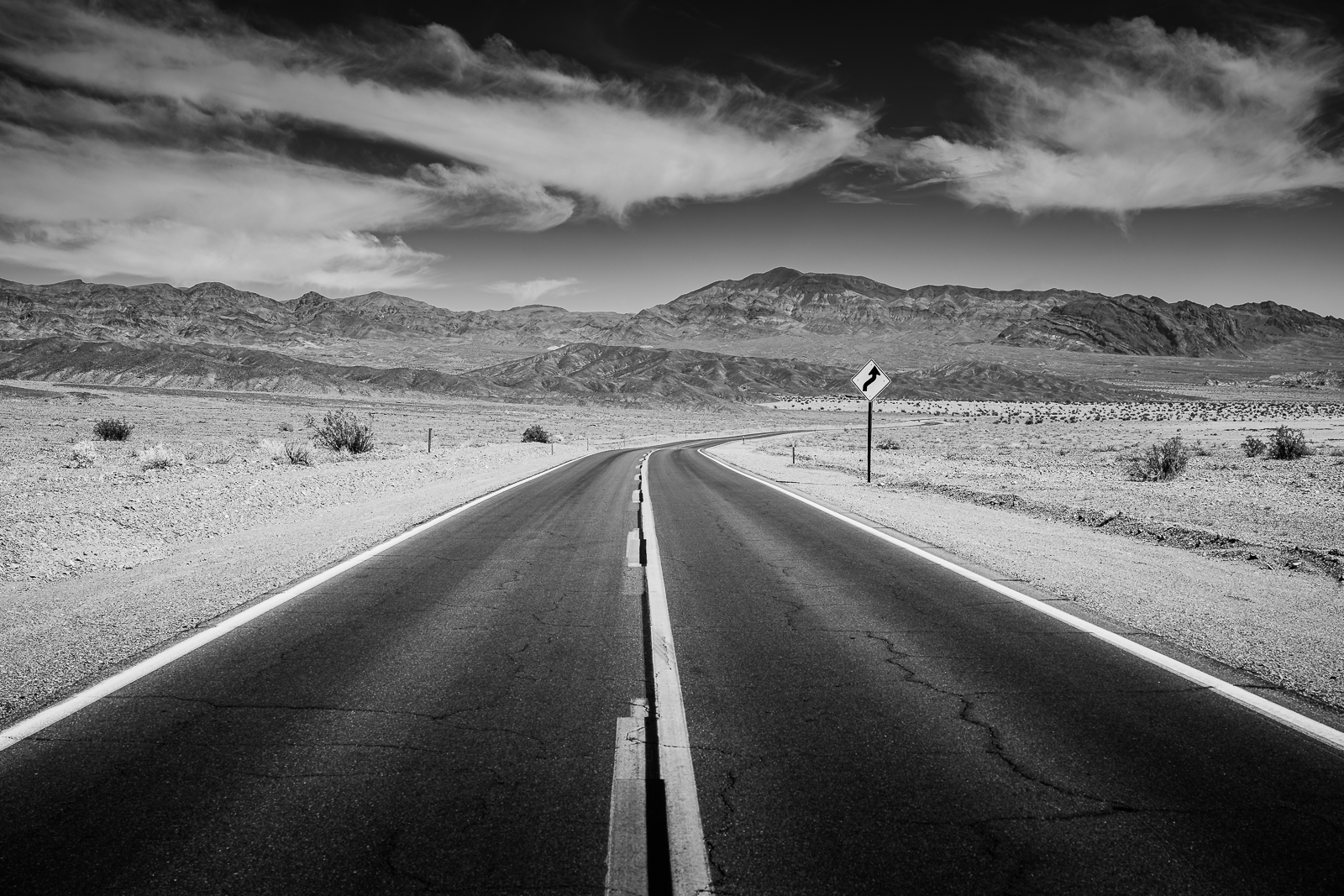 A black-and-white photo of a winding road in a desert landscape, with mountains in the background and scattered bushes, under a sky with wispy clouds.