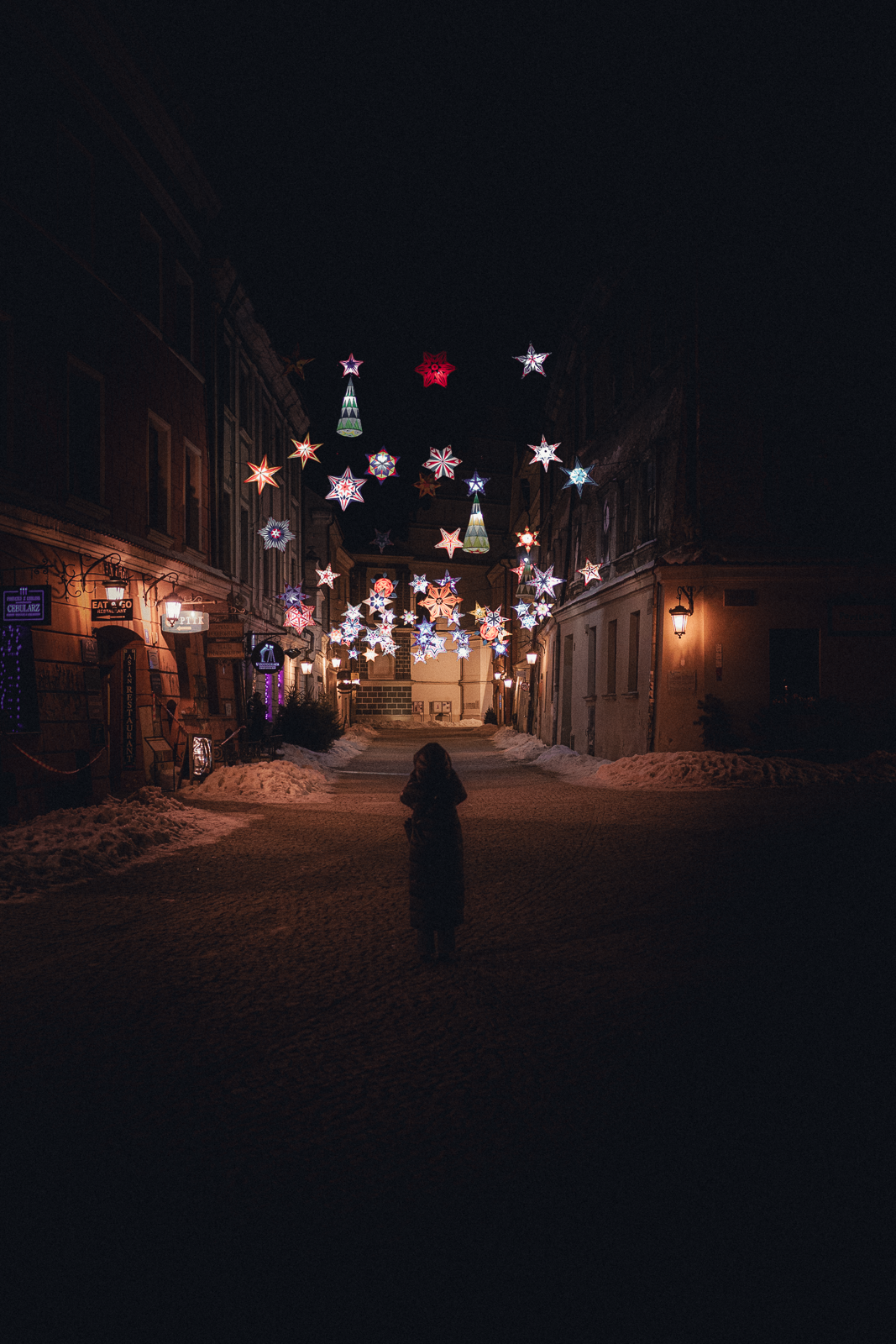 A dark street decorated with colorful, illuminated star-shaped holiday lights hanging above. A person stands in the middle of the street, facing the lights, with snow on the sides of the street.