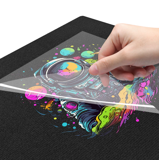 A hand peeling clear transfer film off a vibrant, multi-coloured custom DTF printed astronaut design on a black t-shirt. The final step of our premium Direct-to-Film (DTF) printing process. DTF is incredibly vibrant, durable, and stretch-resistant.