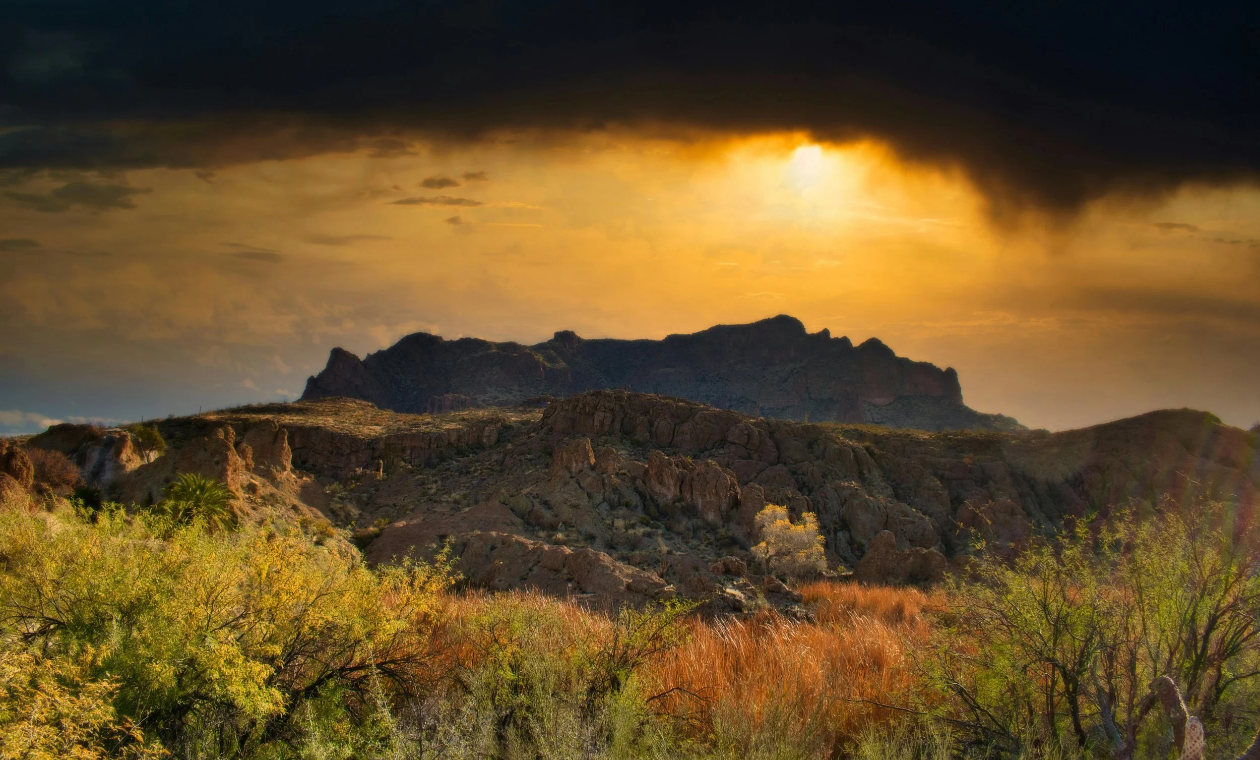 A desert landscape at sunset with rocky hills, sparse vegetation, and dark clouds in the sky, illuminated by a golden glow.