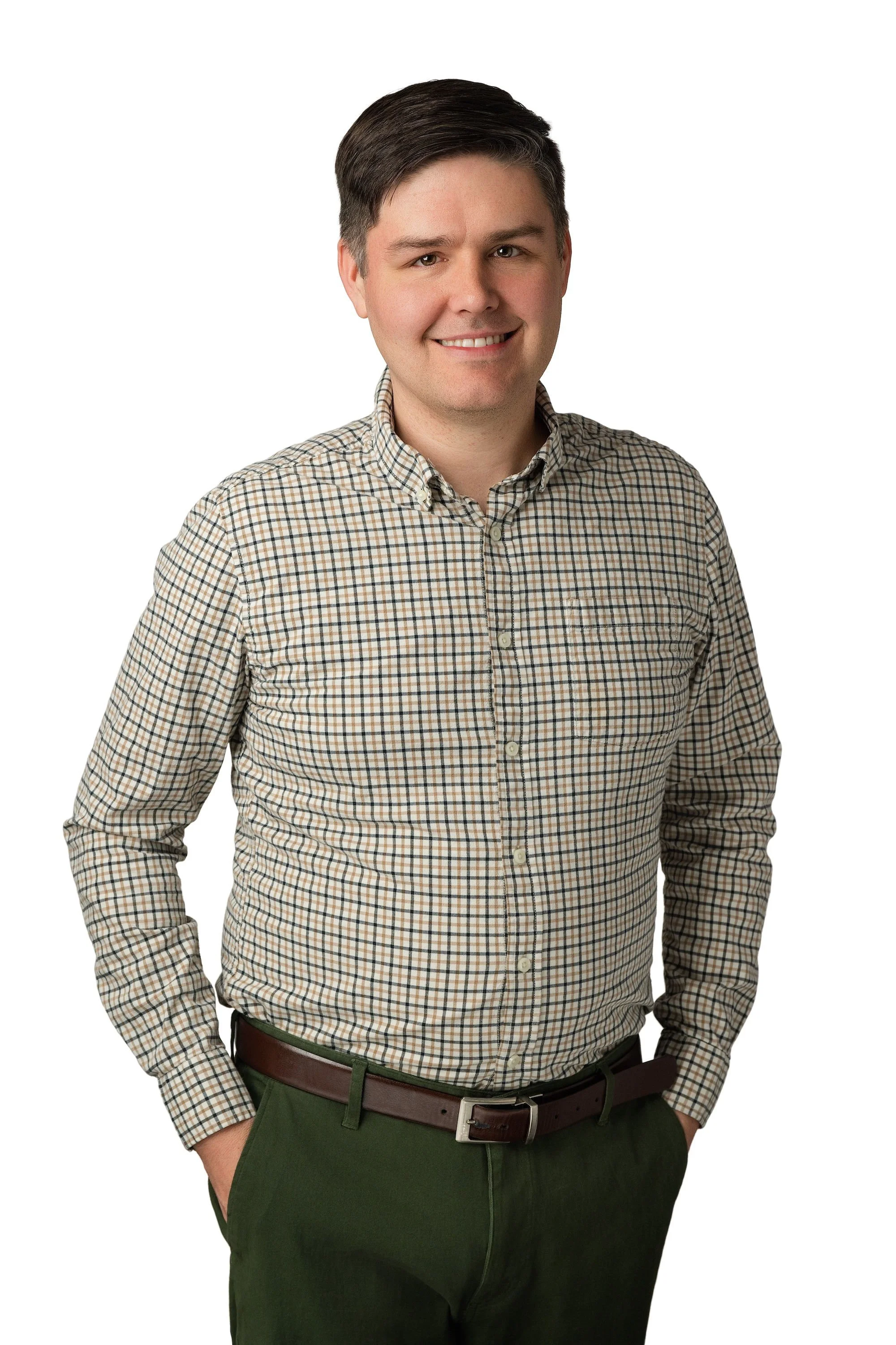 A portrait of a young man with dark hair, wearing a checked shirt and green pants, standing with hands in pockets against a plain white background.