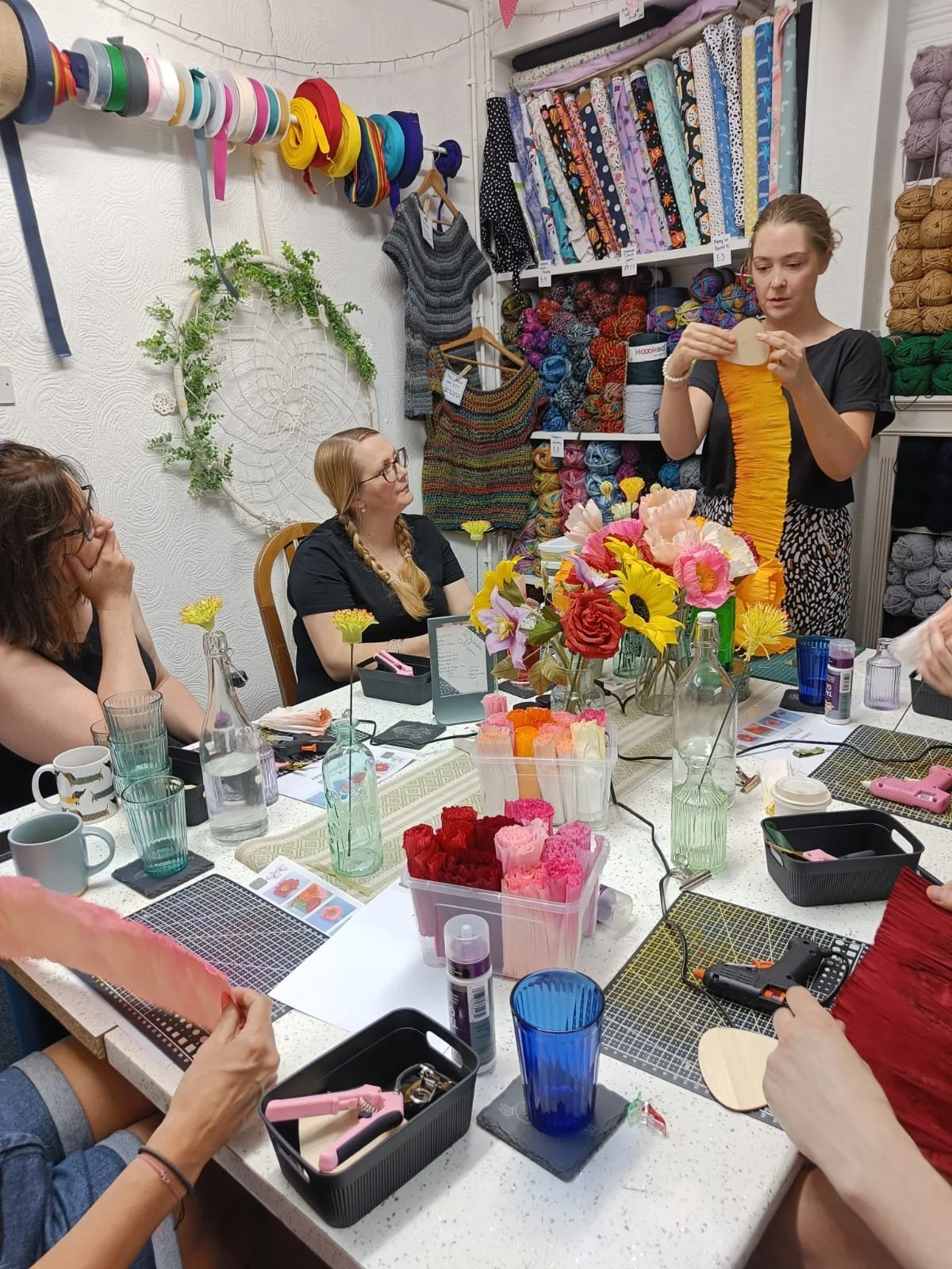 A woman demonstrating flower making with tissue paper at a craft workshop, while seated women watch and craft supplies and colorful flowers are on the table.