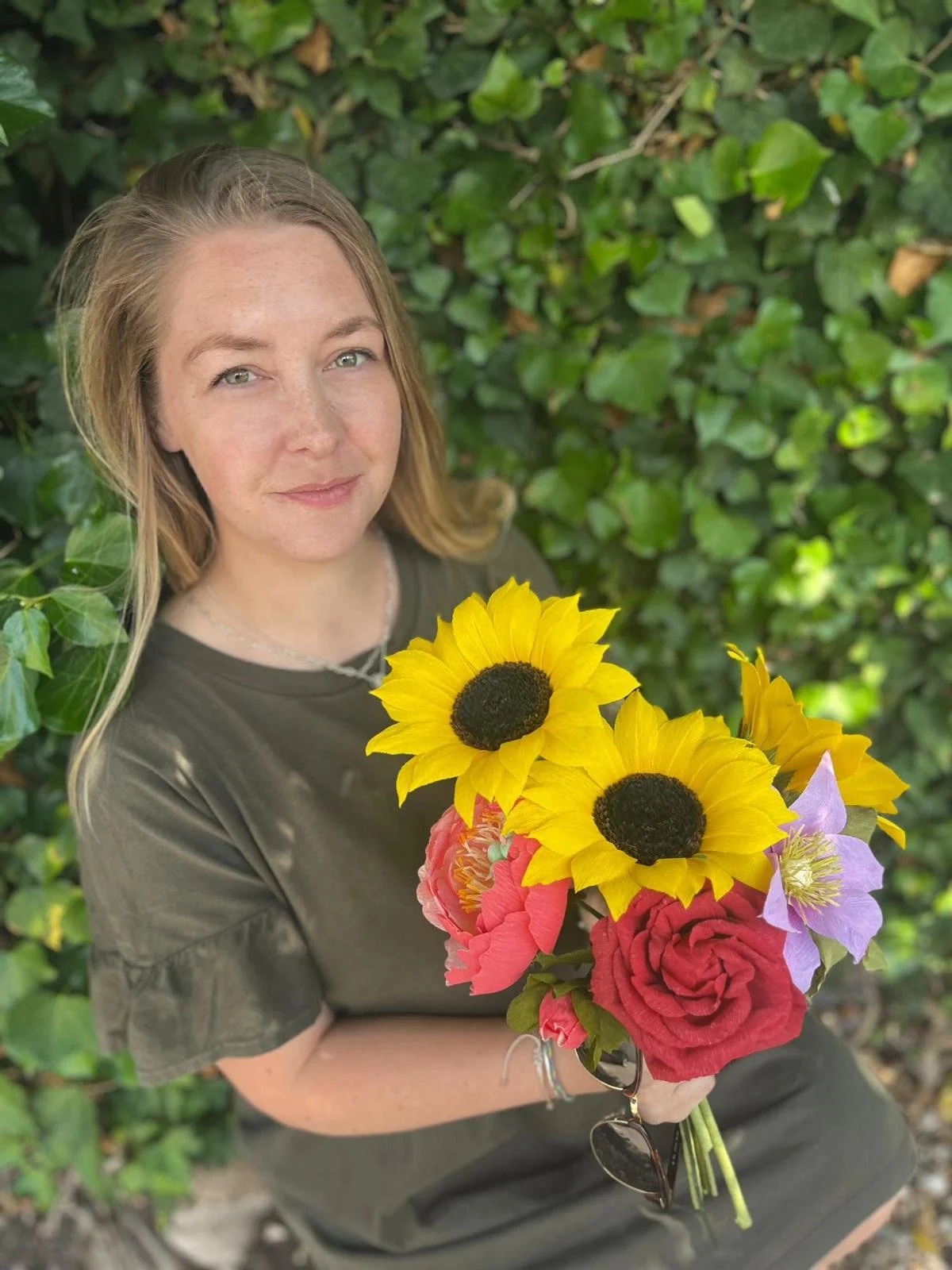 A woman with long blonde hair holding a bouquet of colorful flowers including sunflowers, roses, and other blooms, standing in front of green leafy bushes.