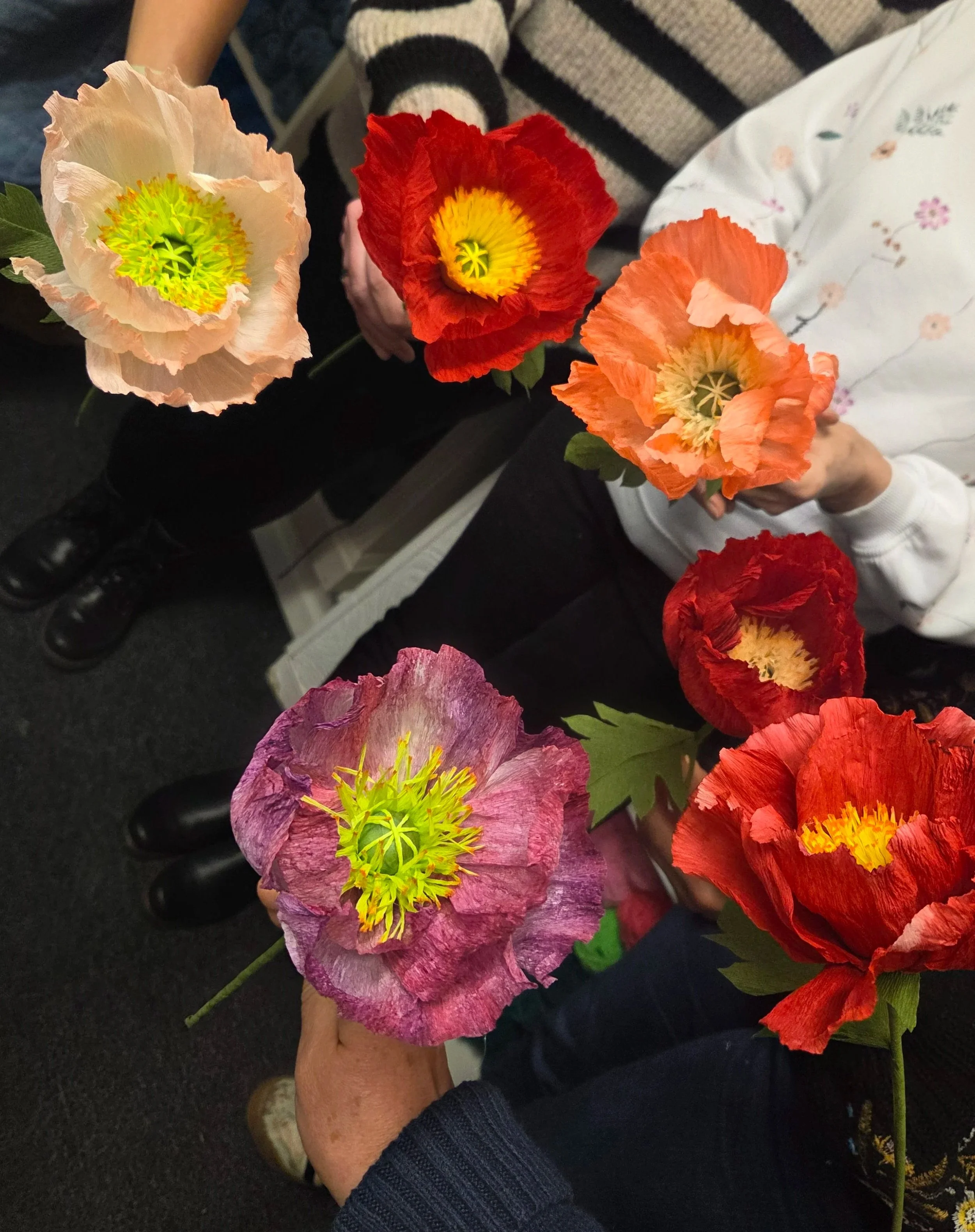 People holding colorful artificial poppy flowers in various shades of pink, red, purple, and orange.