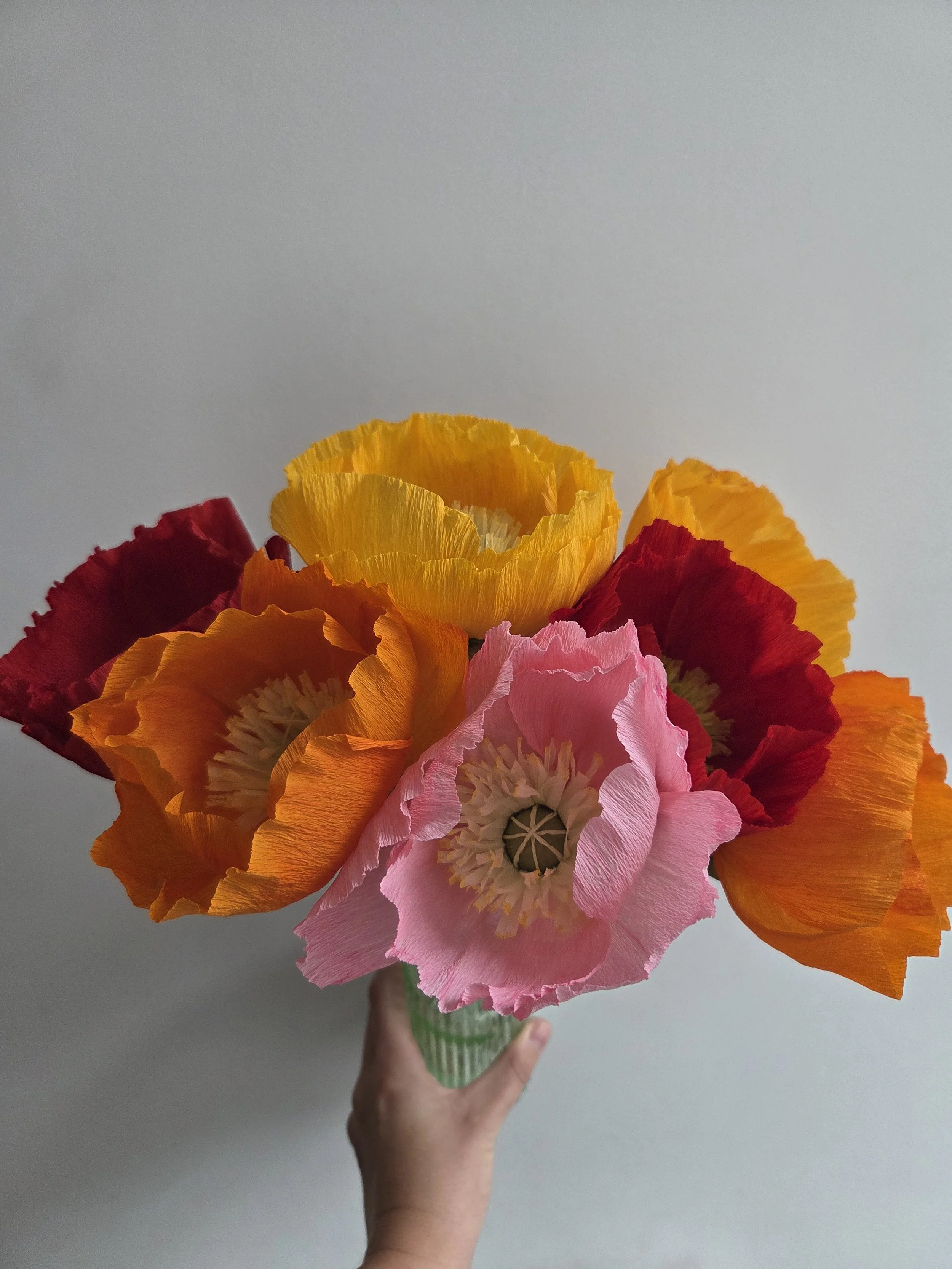 Hand holding a colorful bouquet of paper poppy flowers in various shades of pink, orange, yellow, and red against a plain gray background.