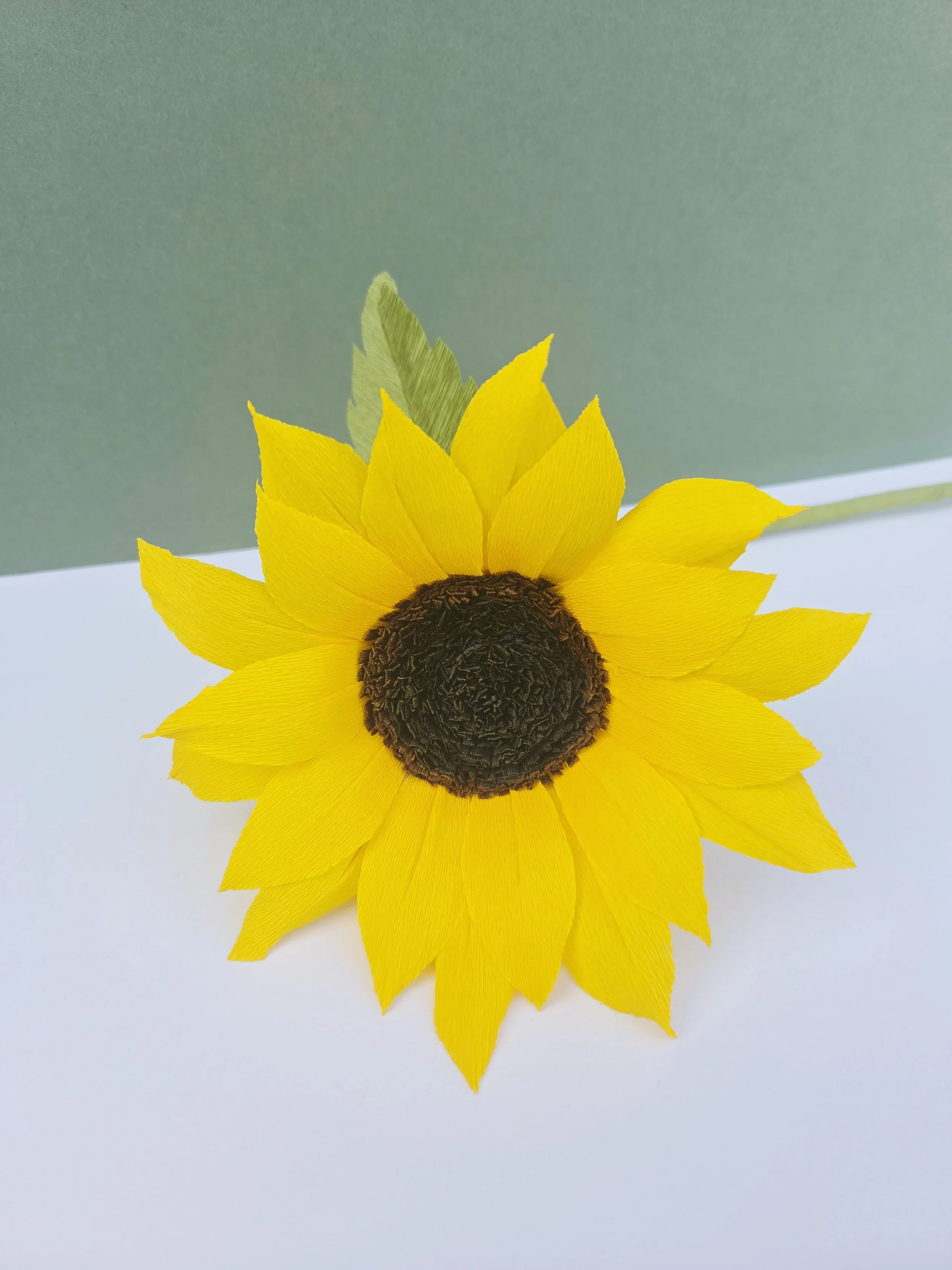 Artificial sunflower with yellow petals, green leaves, and a dark brown center, placed on a white and green background.