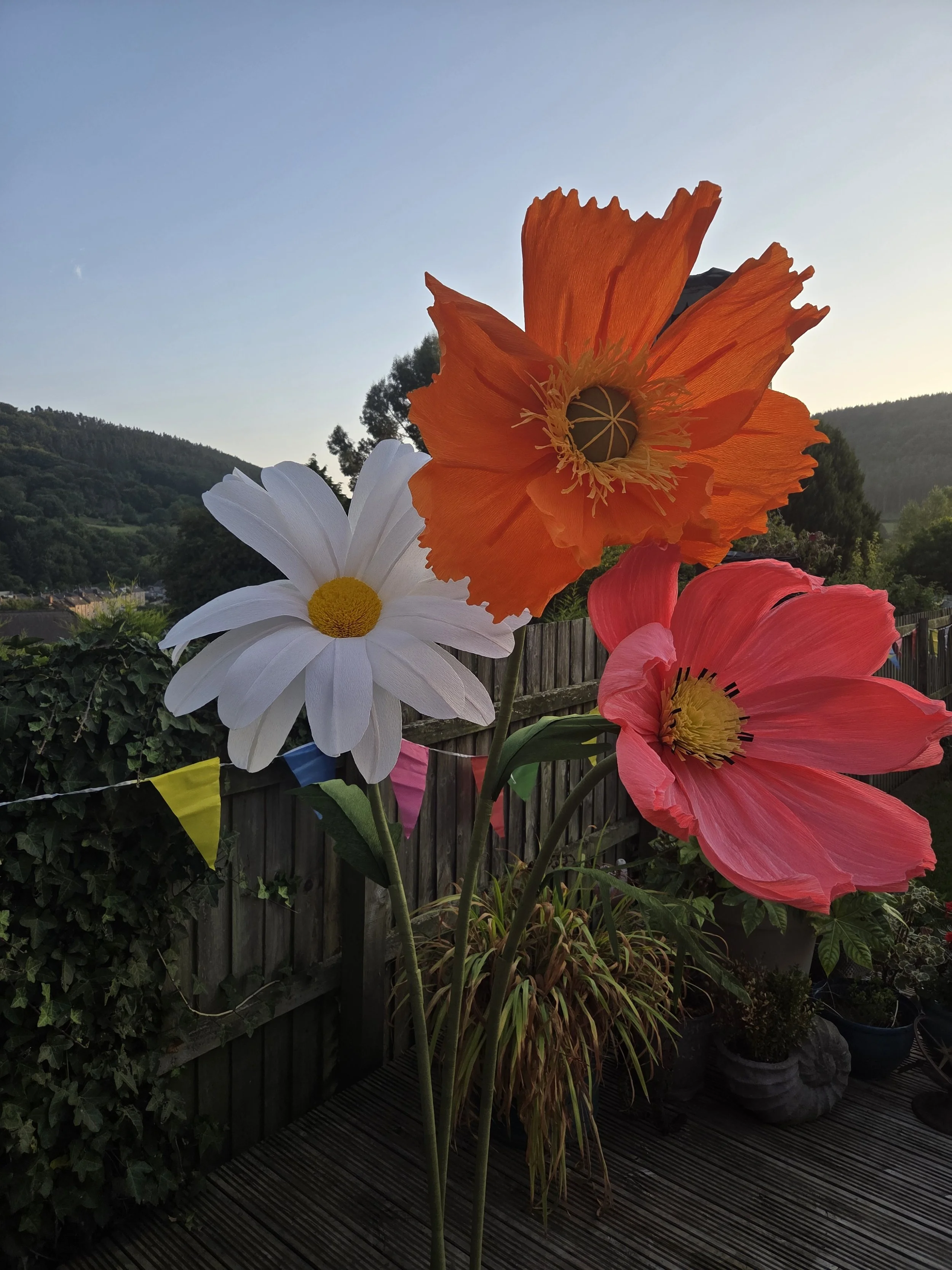 Close-up of three large artificial flowers, orange, white, and pink, on tall stems outdoors with a wooden fence, potted plants, and a scenic hilly background.