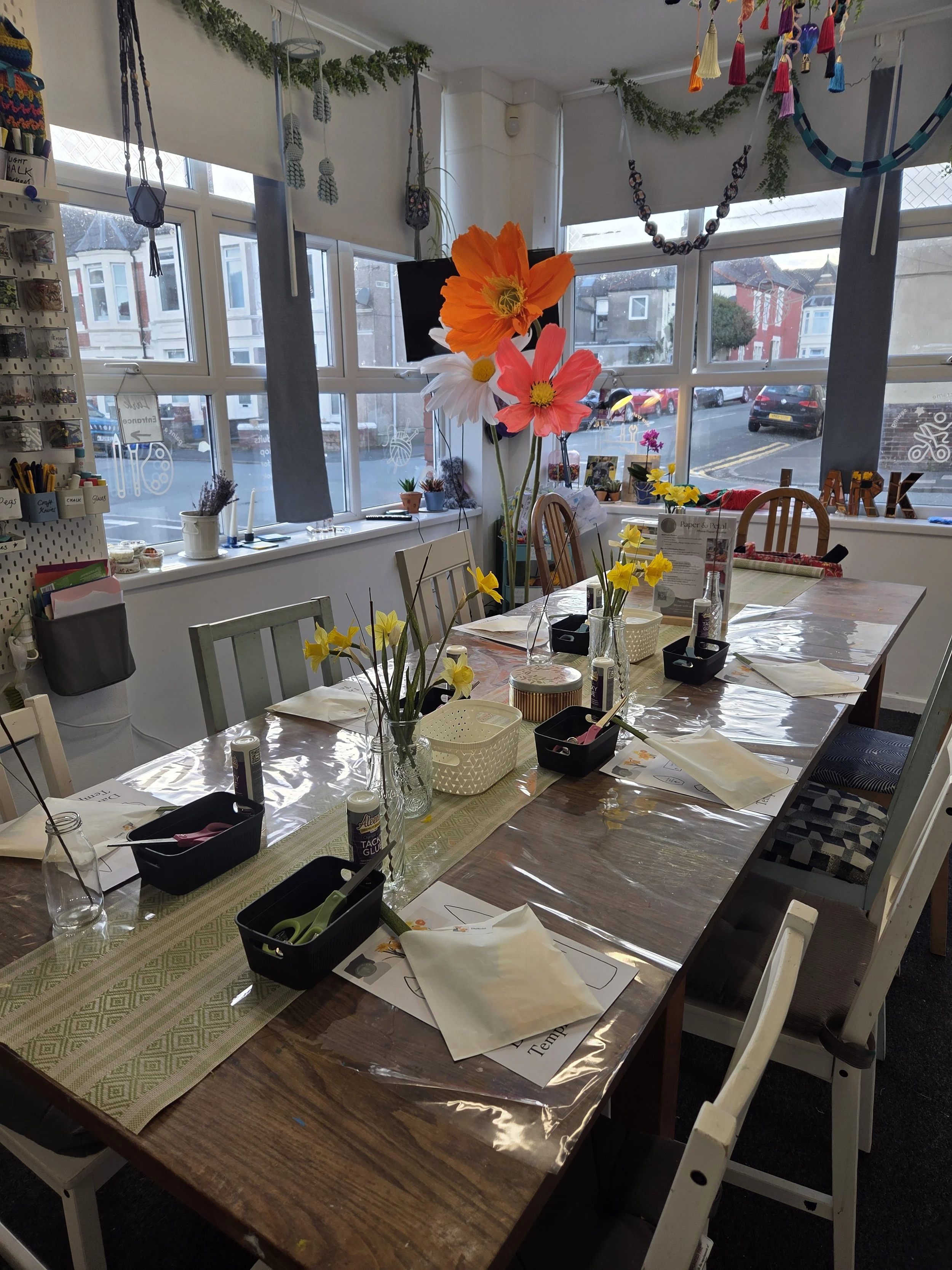 A decorated craft or workshop space with a long table, flowers, and supplies for an activity. The table has a green table runner and is set with papers, scissors, glue, and napkins. Large paper flowers in orange, pink, and white with yellow centers are in vases. The room has large windows with views of buildings and parked cars outside, and colorful tassel and string decorations hanging from the ceiling.