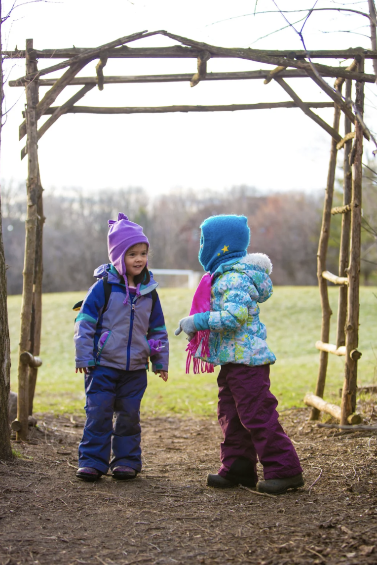 Two kids in kindergarten at Rudolf Steiner School of Ann Arbor are walking through an outdoor wooden archway, wearing warm winter clothes.