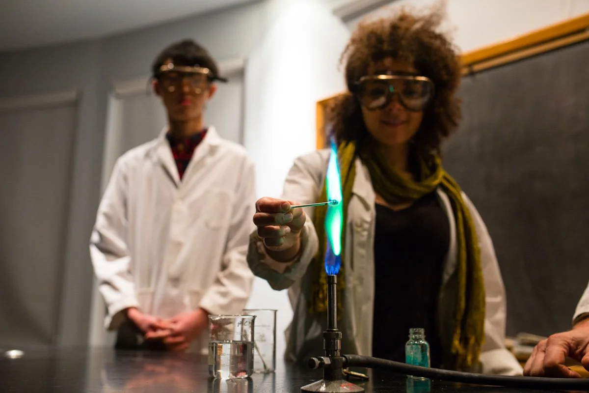 A photo of two students, a boy and a girl, wearing lab coats and safety goggles at Rudolf Steiner School of Ann Arbor during science class. The girl is doing an experiment with a flame while the boy looks on.