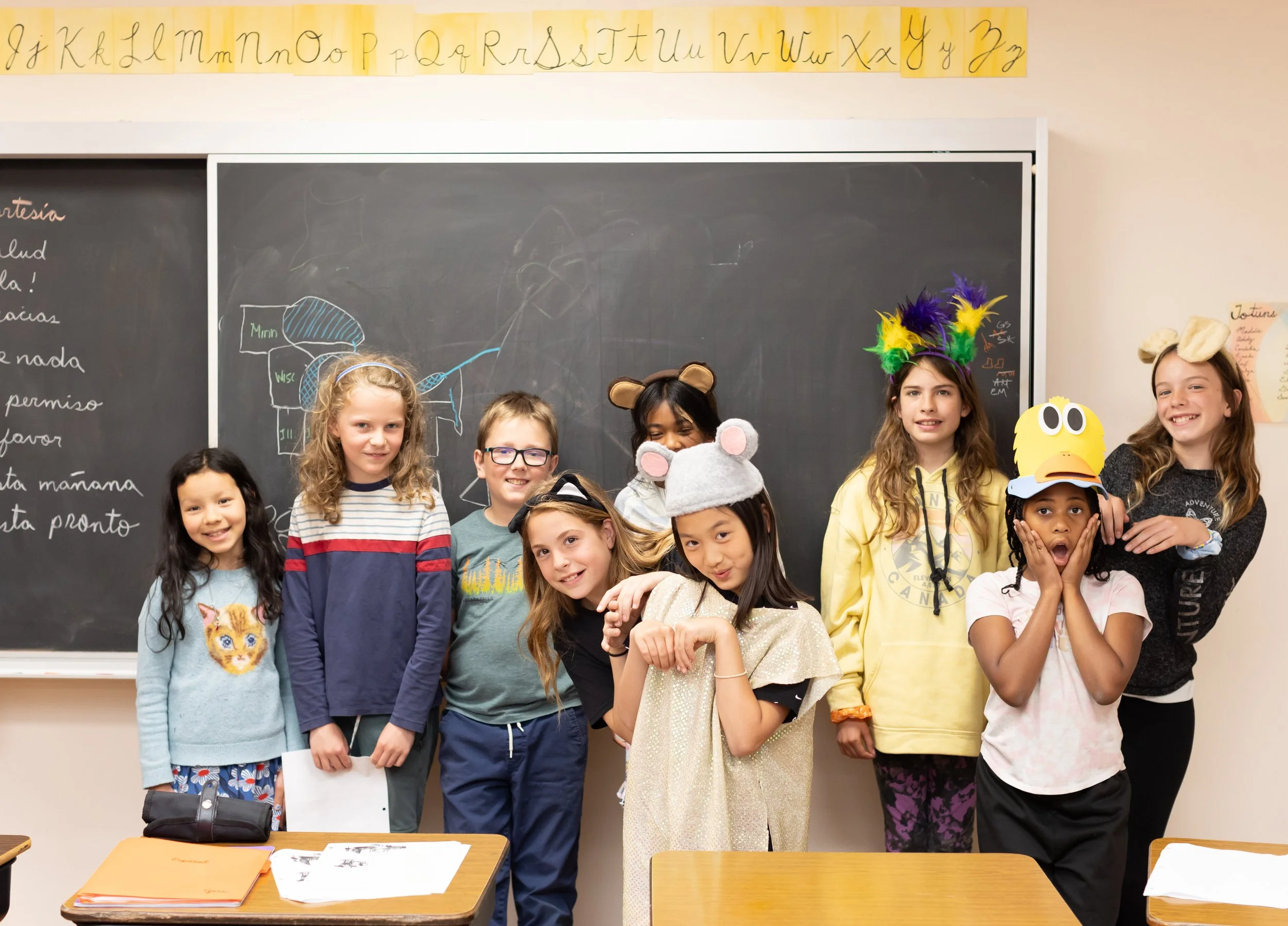 A group of smiling students in elementary school at Rudolf Steiner School of Ann Arbor pose for a silly photo in front of the blackboard in the classroom. Most are wearing a hat with ears or feathers.