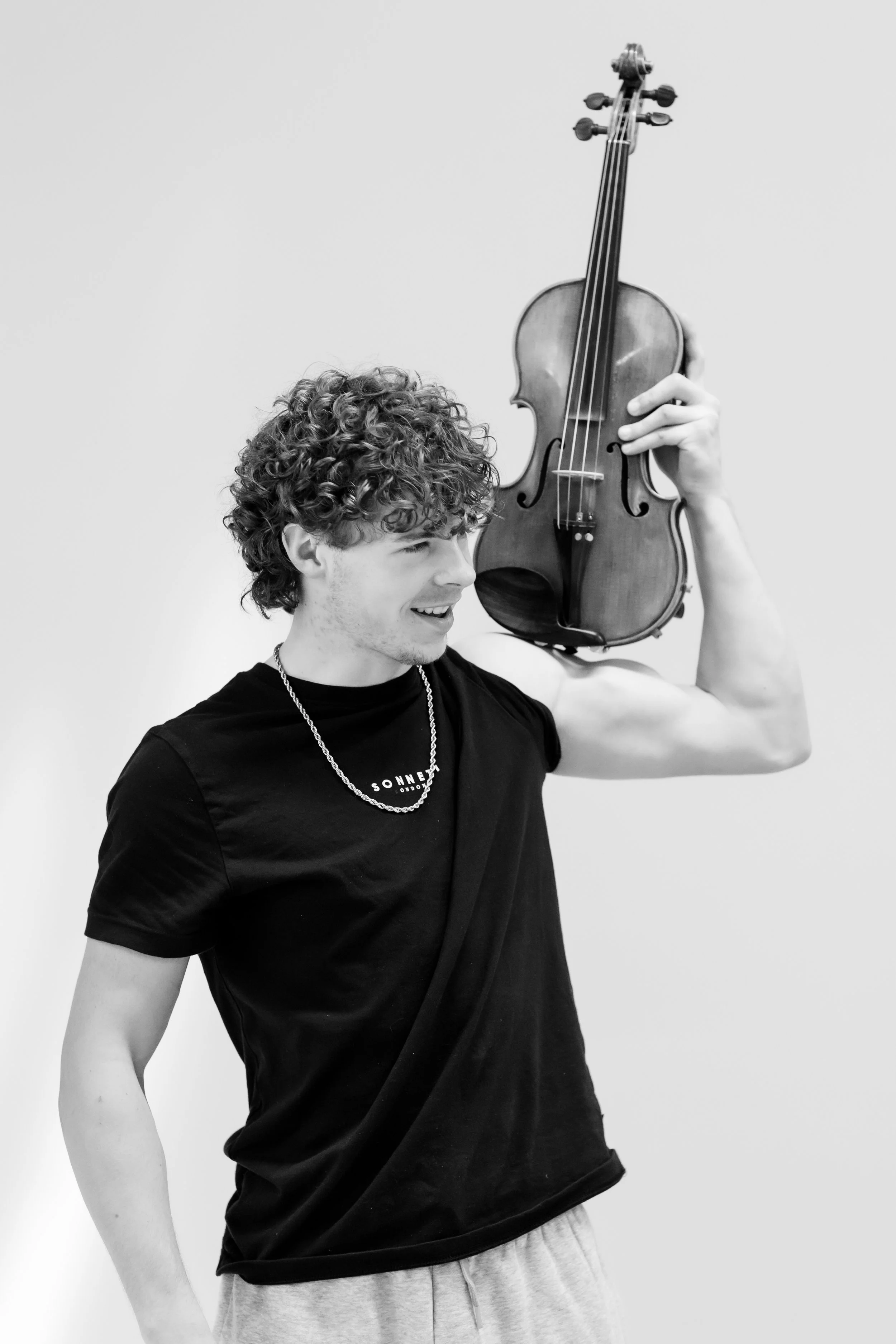 A black and white image of a smiling curly haired boy at Rudolf Steiner School of Ann Arbor. He is holding a violin on his shoulder.