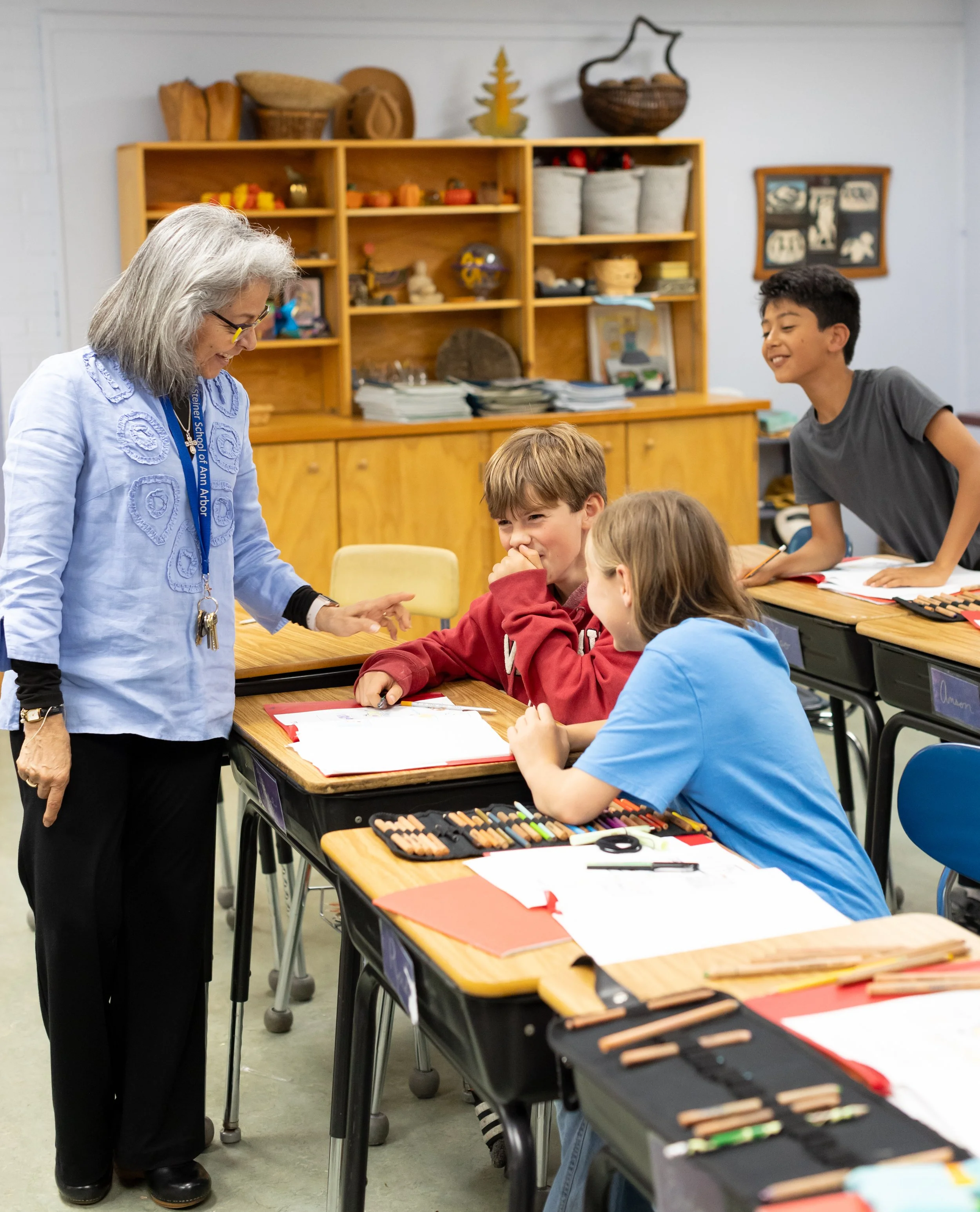 An elementary school teacher at the Rudolf Steiner School of Ann Arbor is looking over a students work. There are 3 kids in the picture, smiling and listening to the teacher.