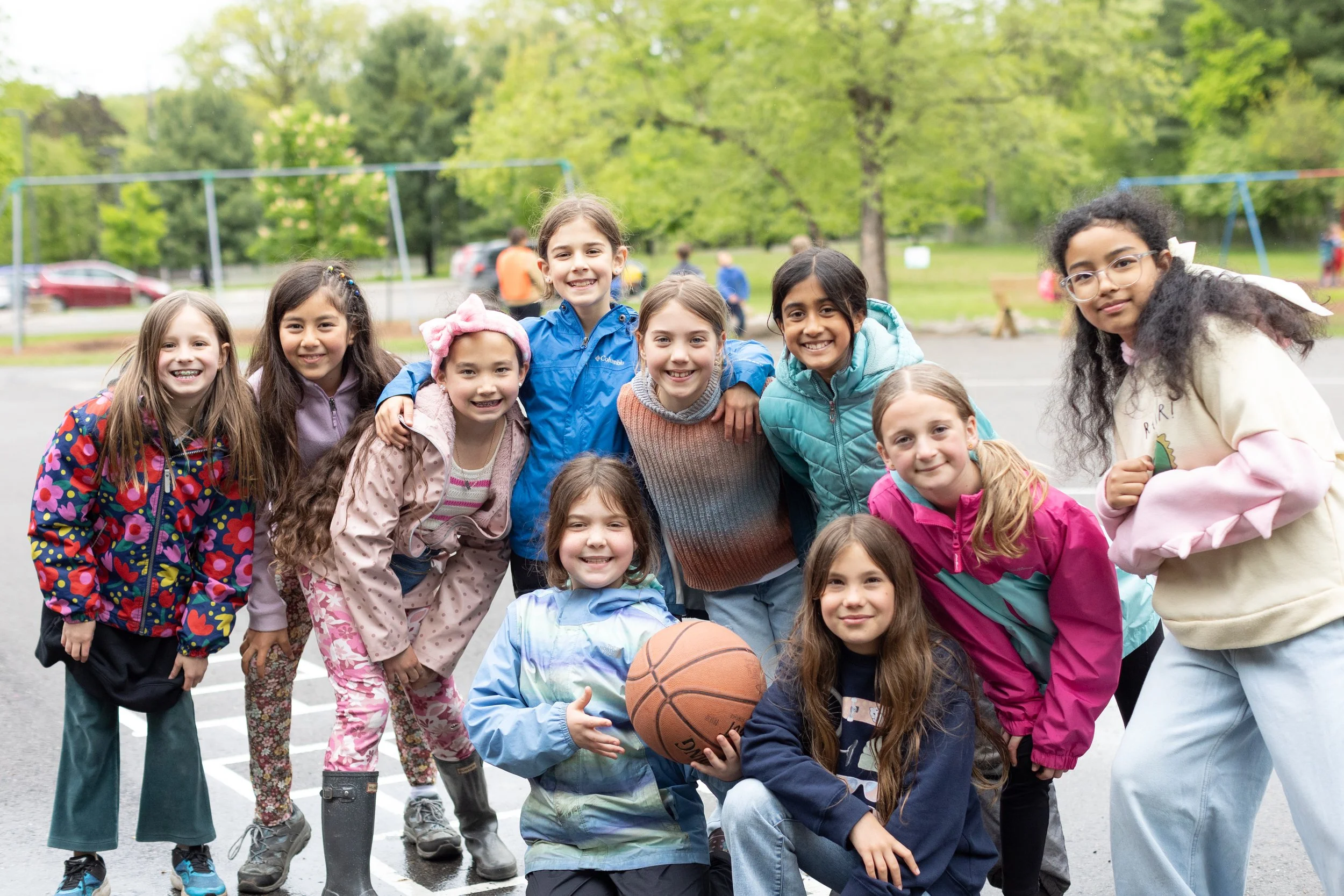 A group of elementary school girls at the Rudolf Steiner School of Ann Arbor are posing for a group photo with a basketball and smiling at the camera.