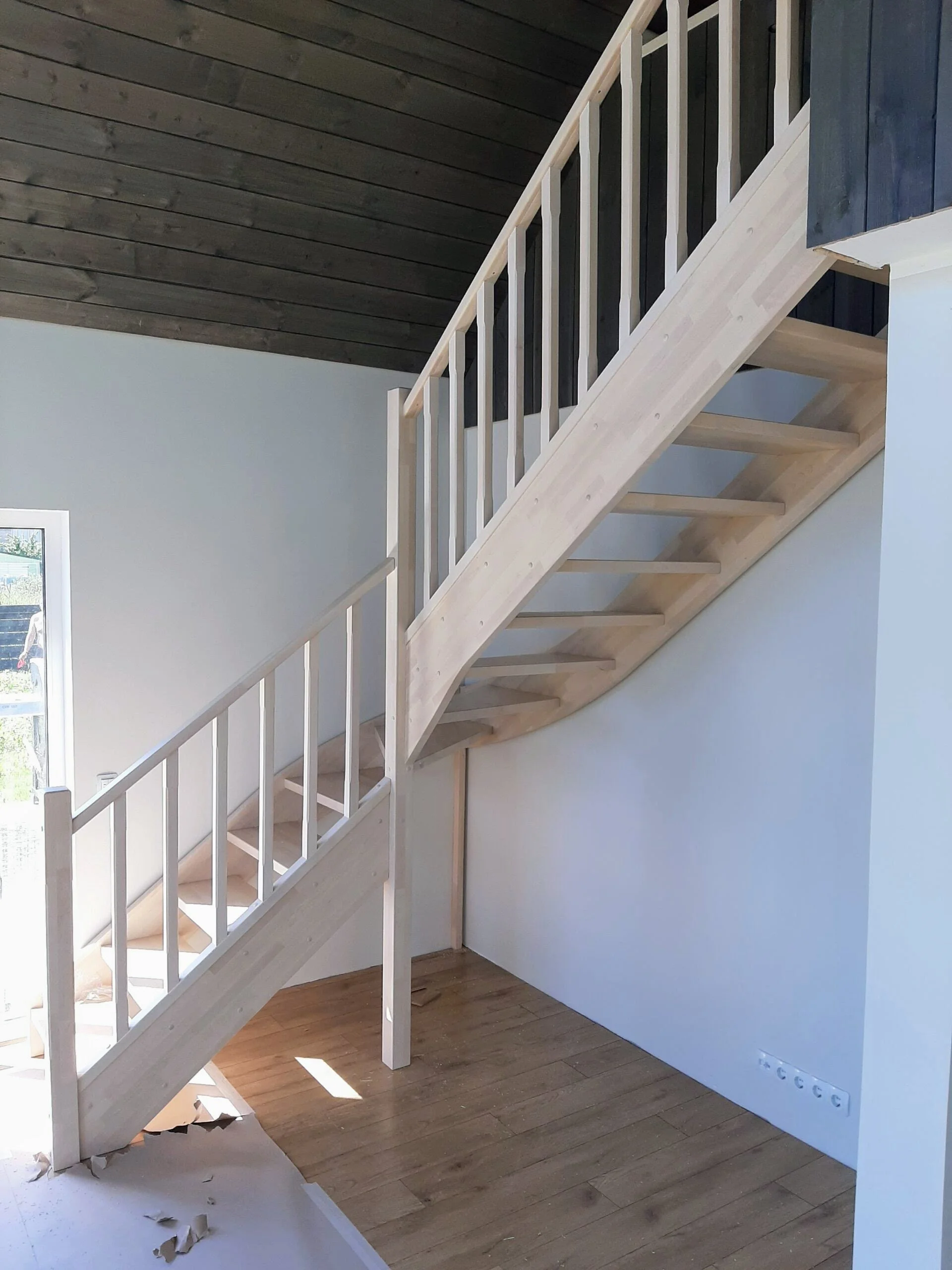 Interior of a house under construction featuring a newly installed wooden staircase with white railings and a dark wood ceiling.