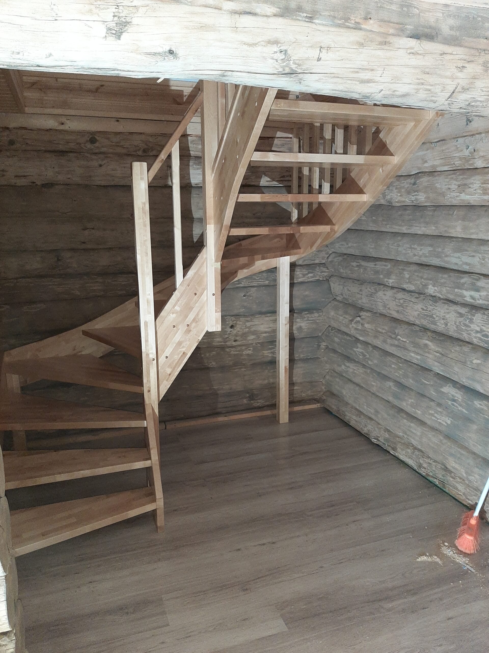 Interior of a wooden staircase with a curved design, installed in a rustic room with log cabin walls and wooden flooring, with a broom leaning against the wall.
