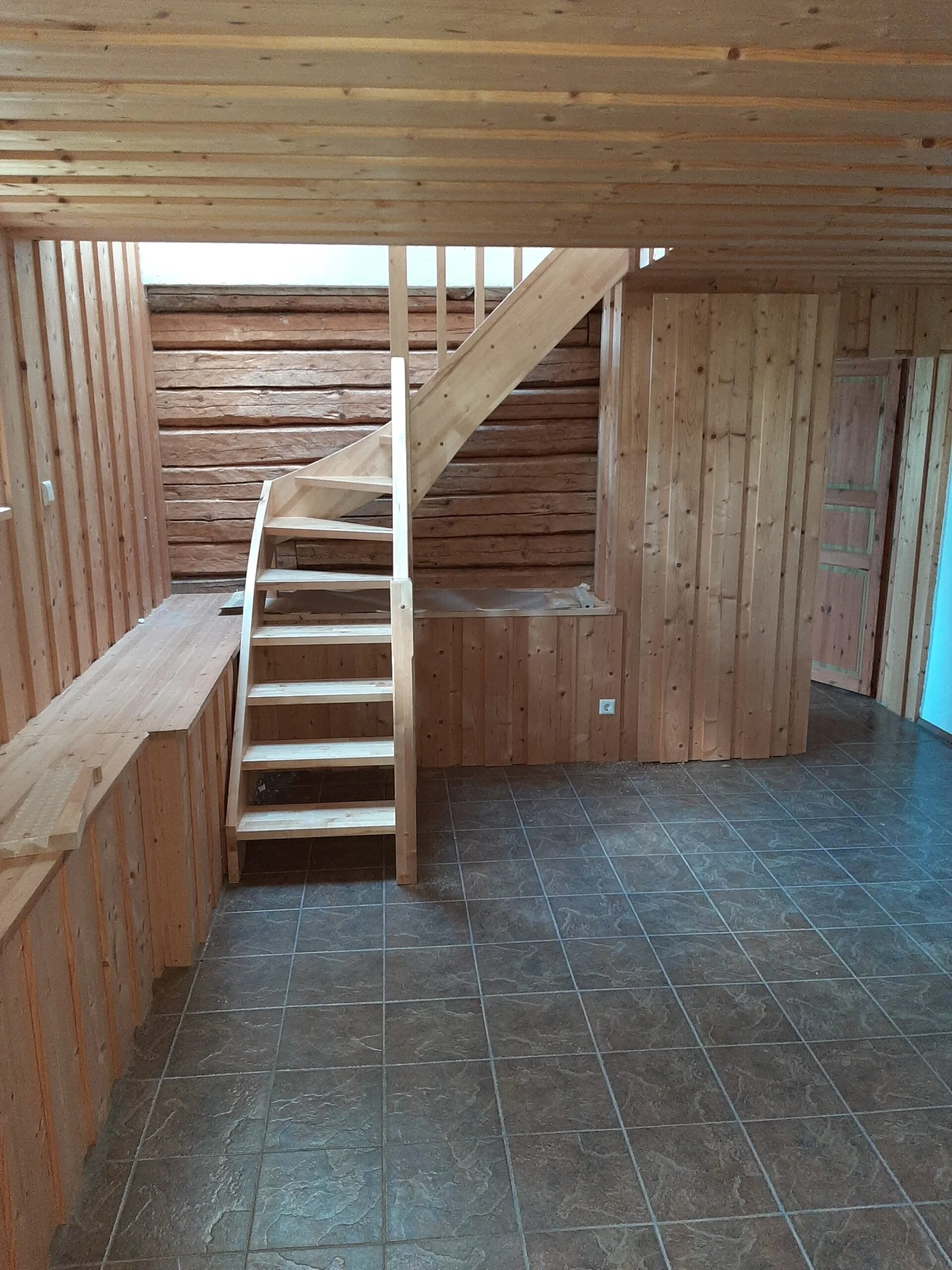 Interior of a wooden cabin with a small staircase leading to an upper level, wood-paneled walls and ceiling, tiled floor, and a small enclosed area on the right.