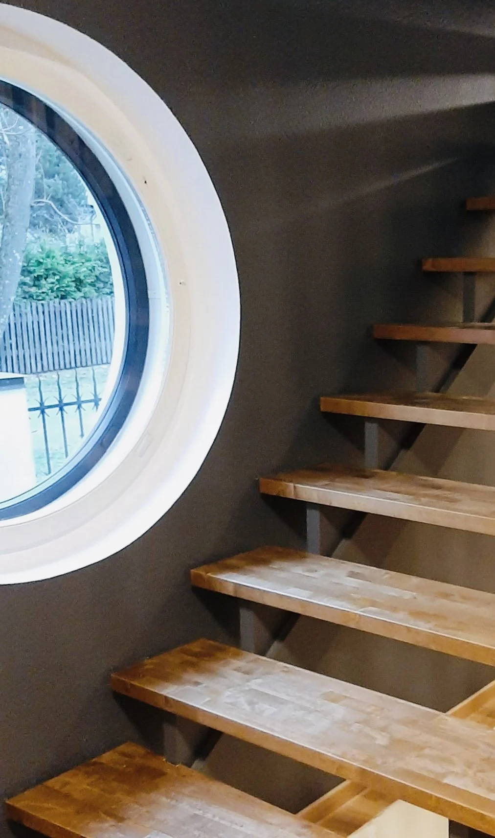 Indoor staircase with wooden steps and a circular window with a view of trees and a fence outside.