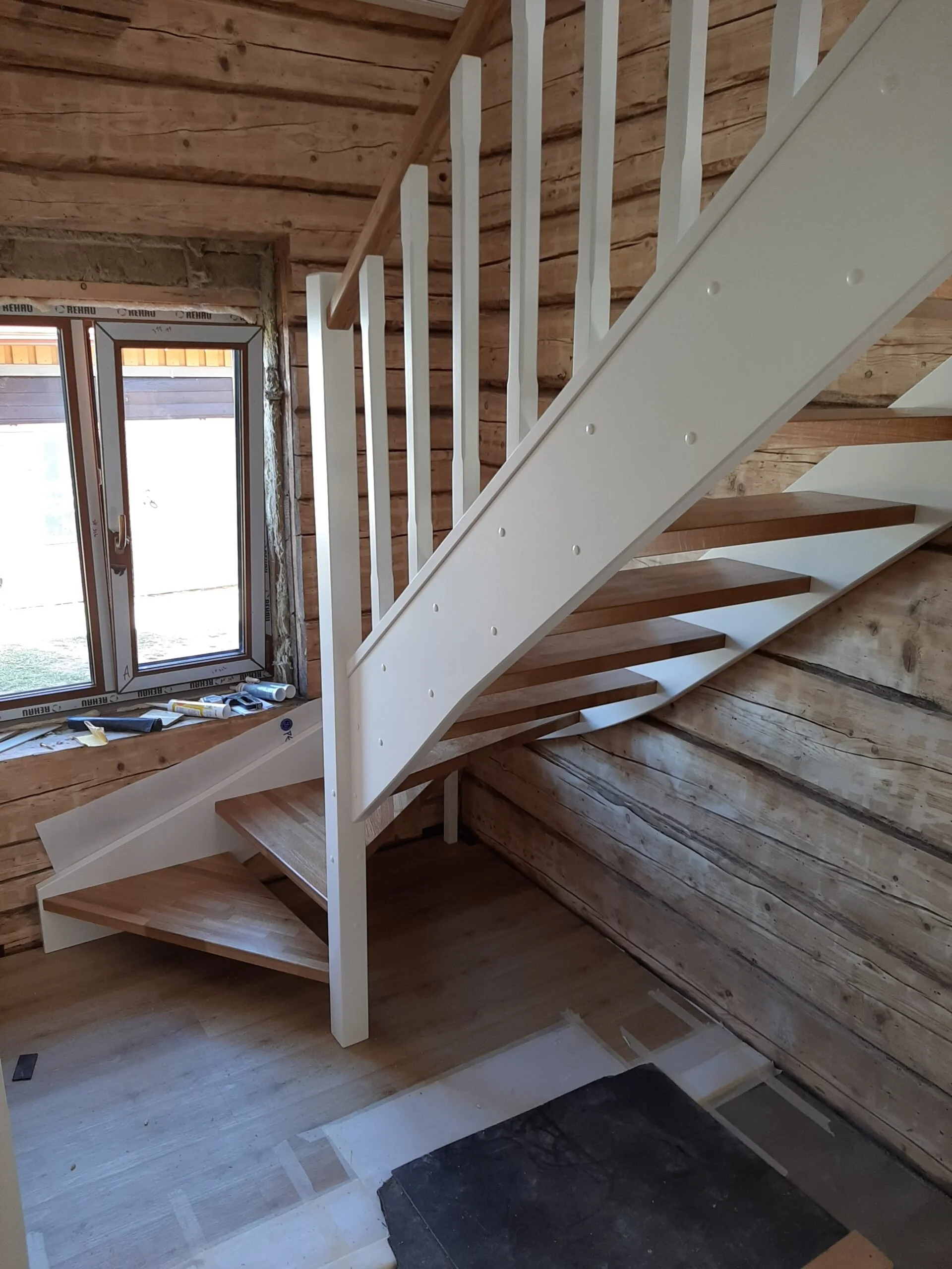 Interior of a wooden house under construction with a staircase that has wooden steps and a white railing, next to a window with construction tools on the window sill.