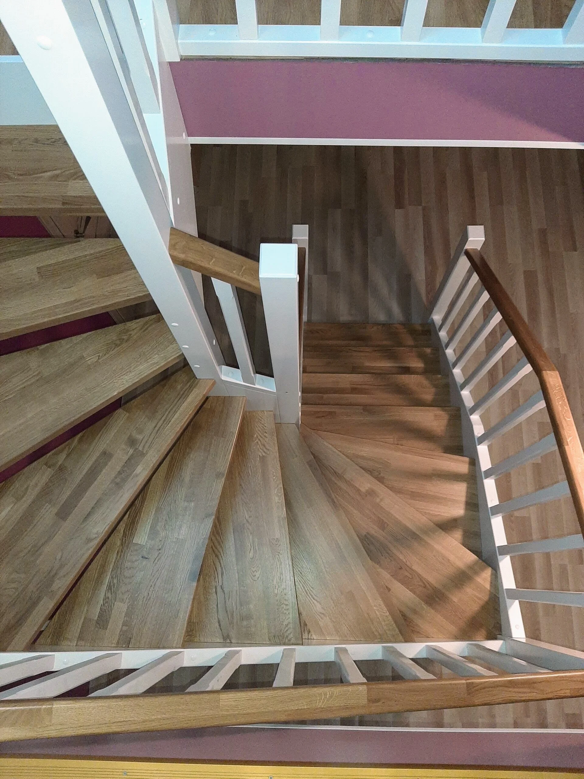 A top-down view of a spiral staircase with wooden steps and white railings, leading down to a lower floor with wood flooring and pink walls.