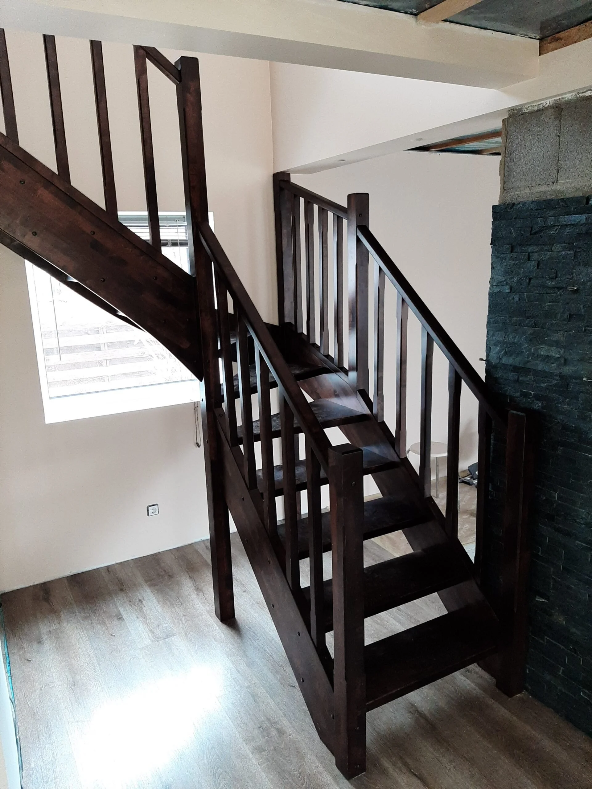 Interior of a house under construction with a dark wooden staircase, a window, and a partially finished wall.