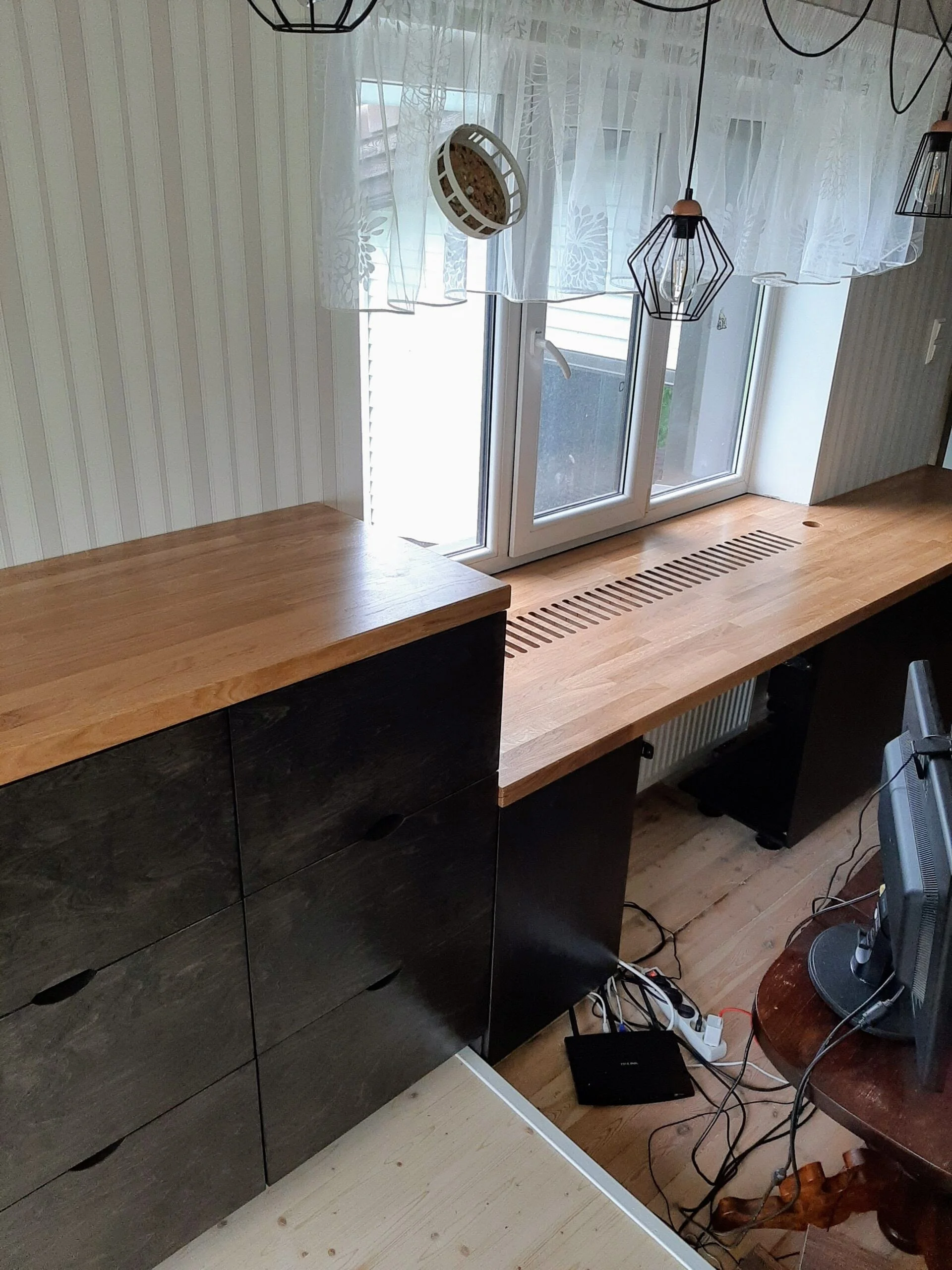 Interior view of a living room with a wooden cabinet, a wooden window sill, and a window with curtains. There are decorative pendant lights hanging from the ceiling, a small television on a wooden stand, and various electronic cords and devices on th