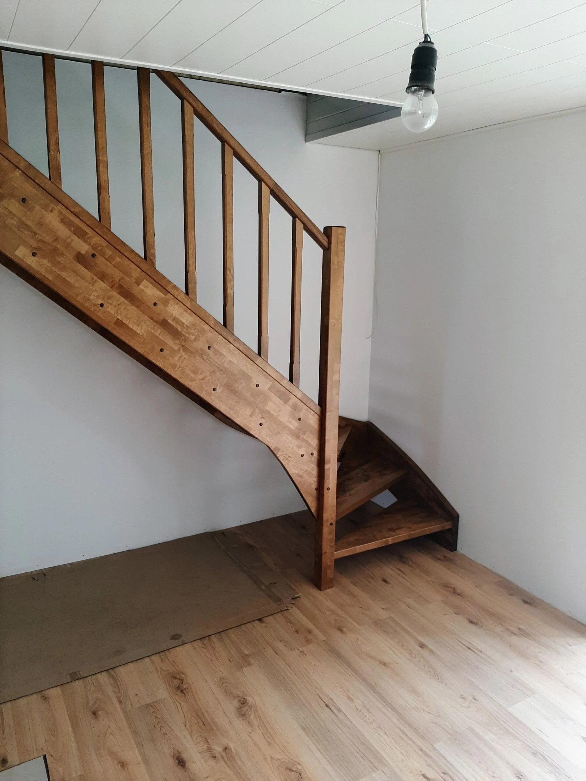 Interior view of a room with a wooden staircase, white walls, a ceiling light bulb, and a wooden floor.