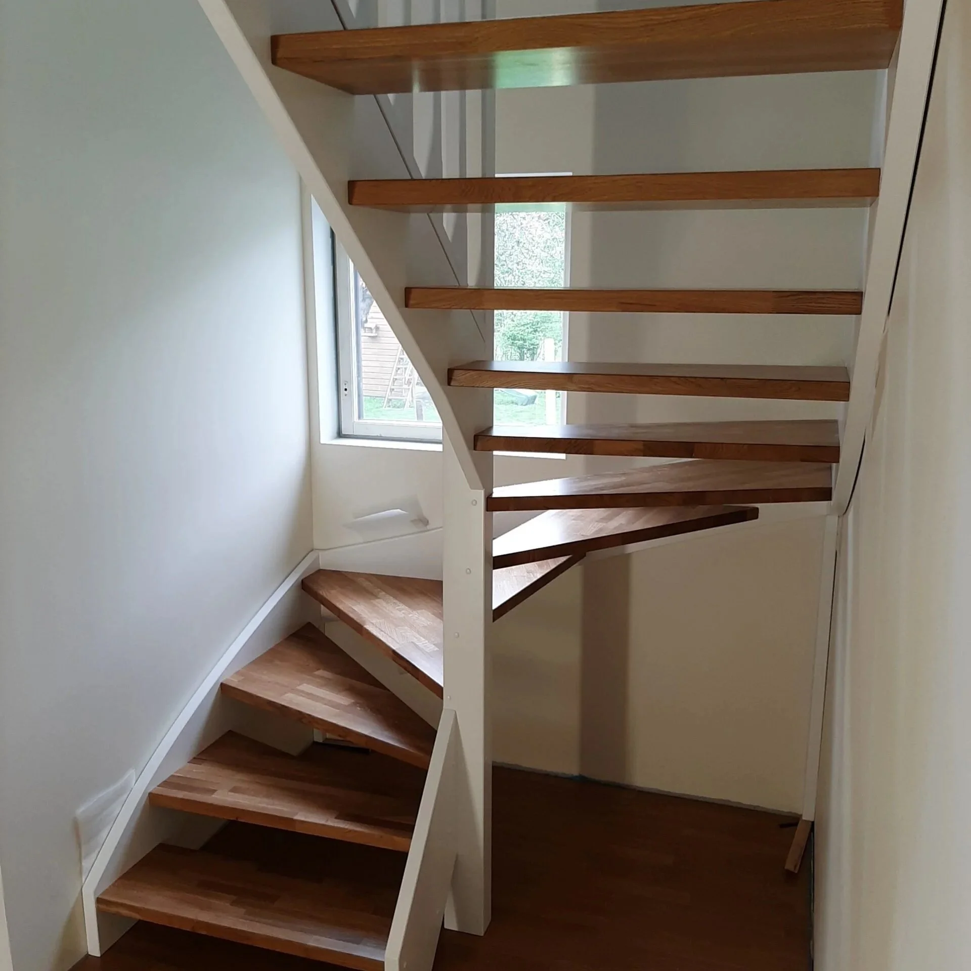 Interior view of a modern staircase with wooden steps and white sides, near a window with a view of a backyard and trees.