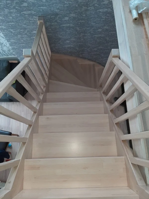 Top-down view of a wooden staircase with light-colored wooden steps and matching railing, leading down to a corner where a textured blue-gray wall and a floor with wood planks meet.